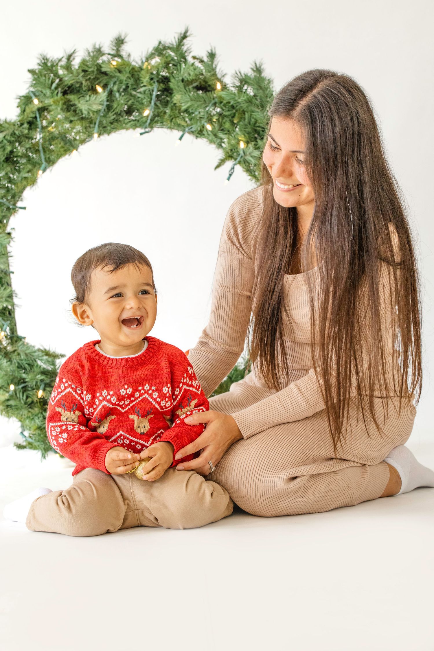 Parent and child in matching holiday outfits sit together against white backdrop with festive greenery decorations.