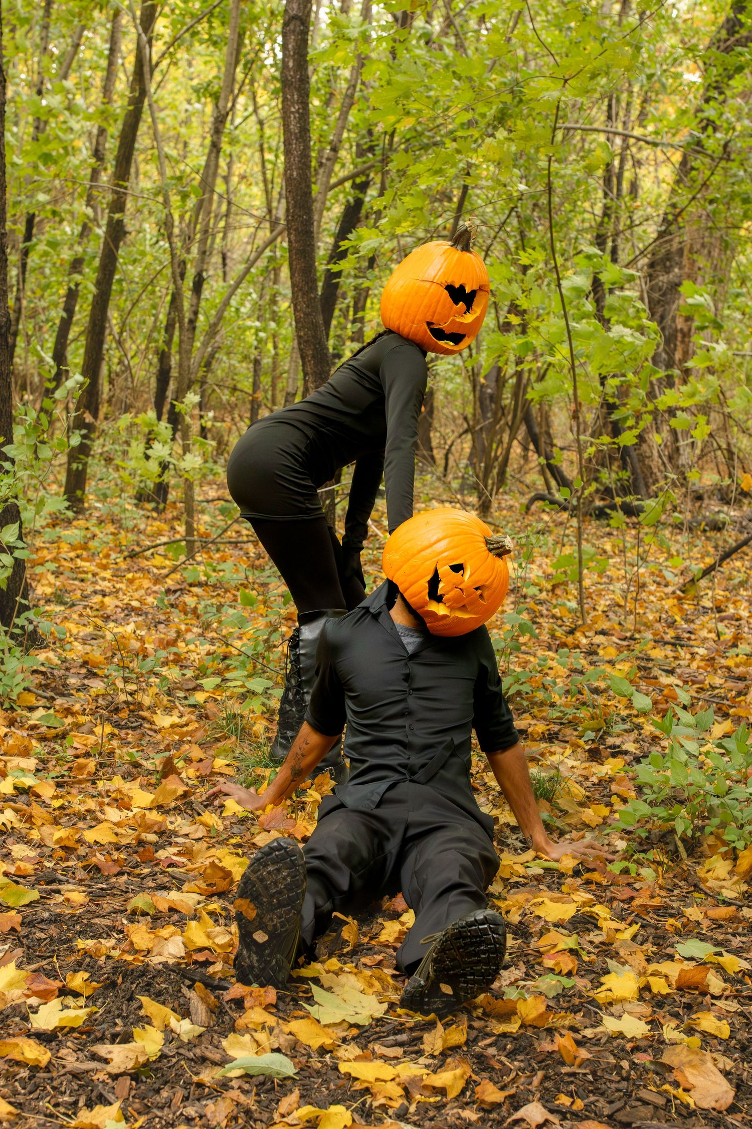 A figure in black clothing wearing a jack-o'-lantern head crawls through autumn leaves in a spooky forest setting.