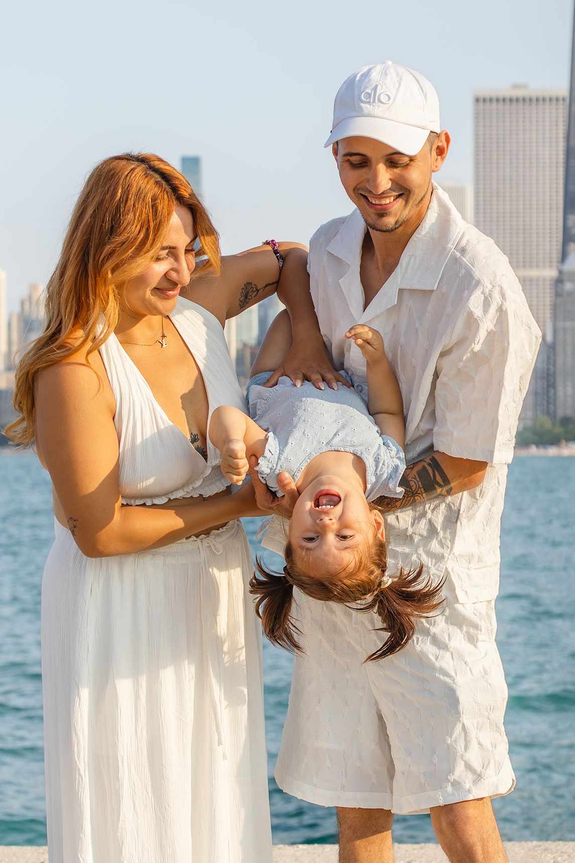 Happy family with young child playing at waterfront, parents in white outfits flipping toddler upside down.