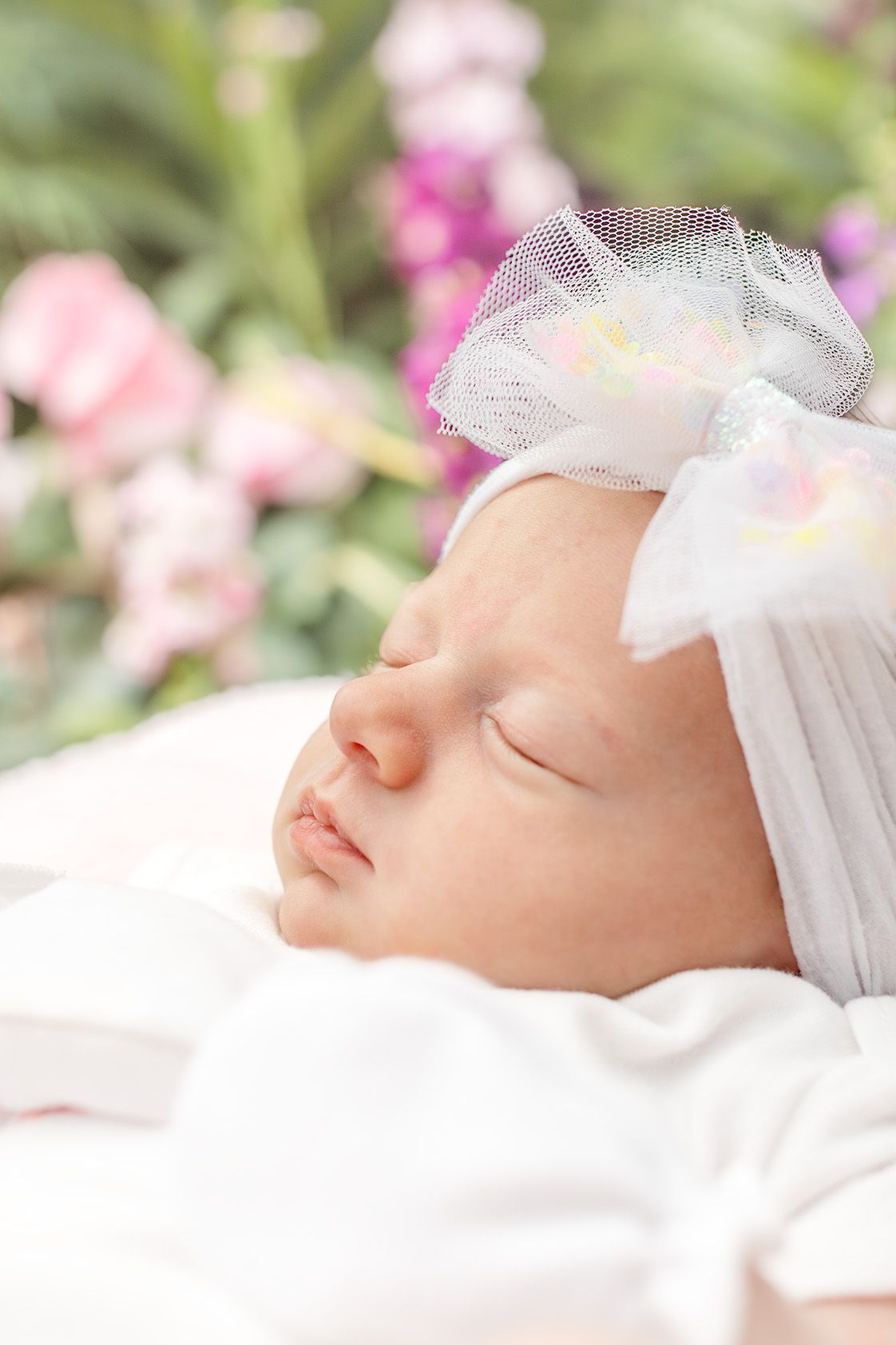 Sleeping newborn baby girl wearing a white headband with bow, surrounded by soft pink and purple flowers.