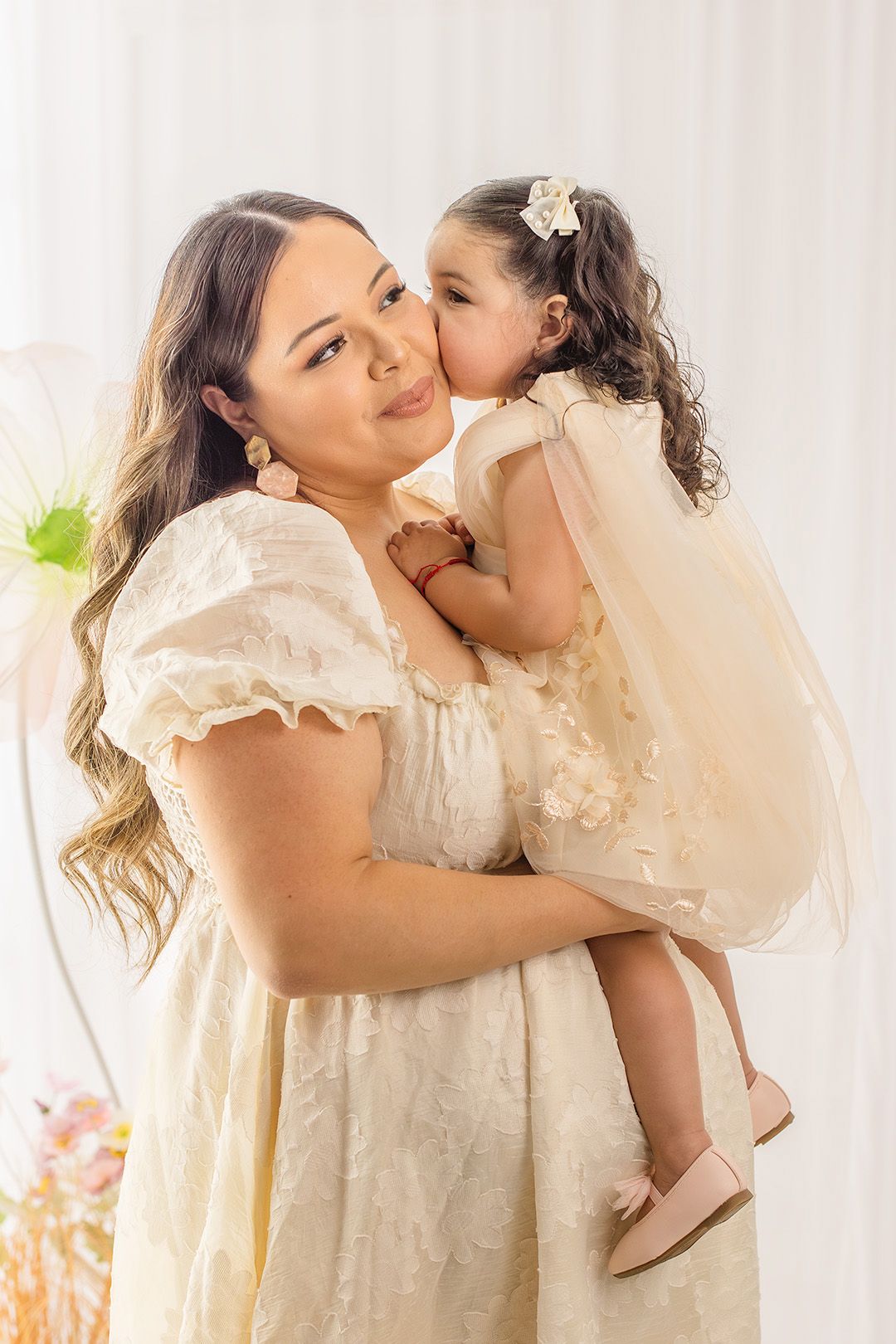 A woman in a cream dress holds a young girl in a matching outfit who kisses her cheek in a bright studio.