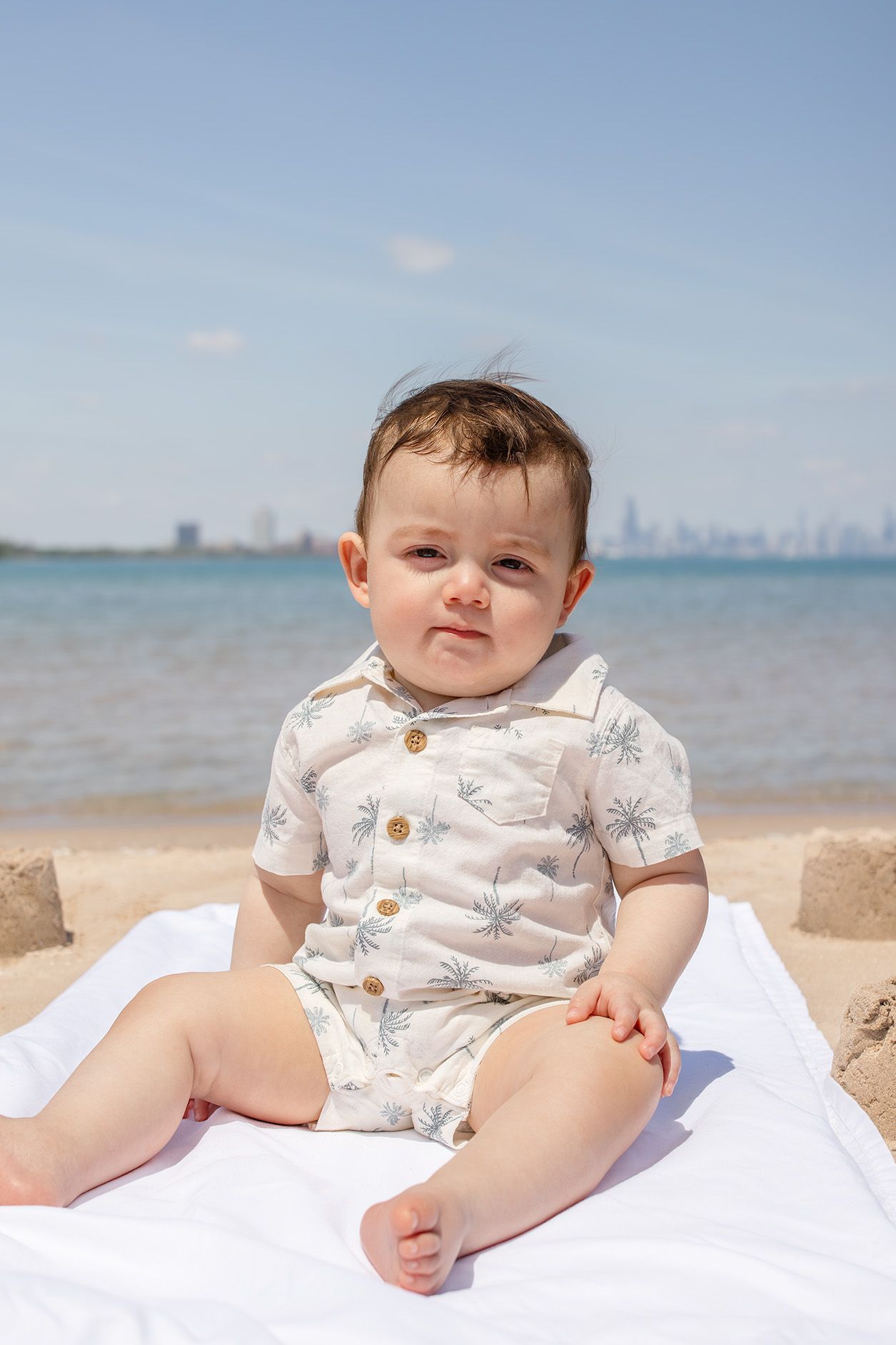Adorable baby sitting on white blanket at sunny beach wearing palm tree romper with city skyline in background.