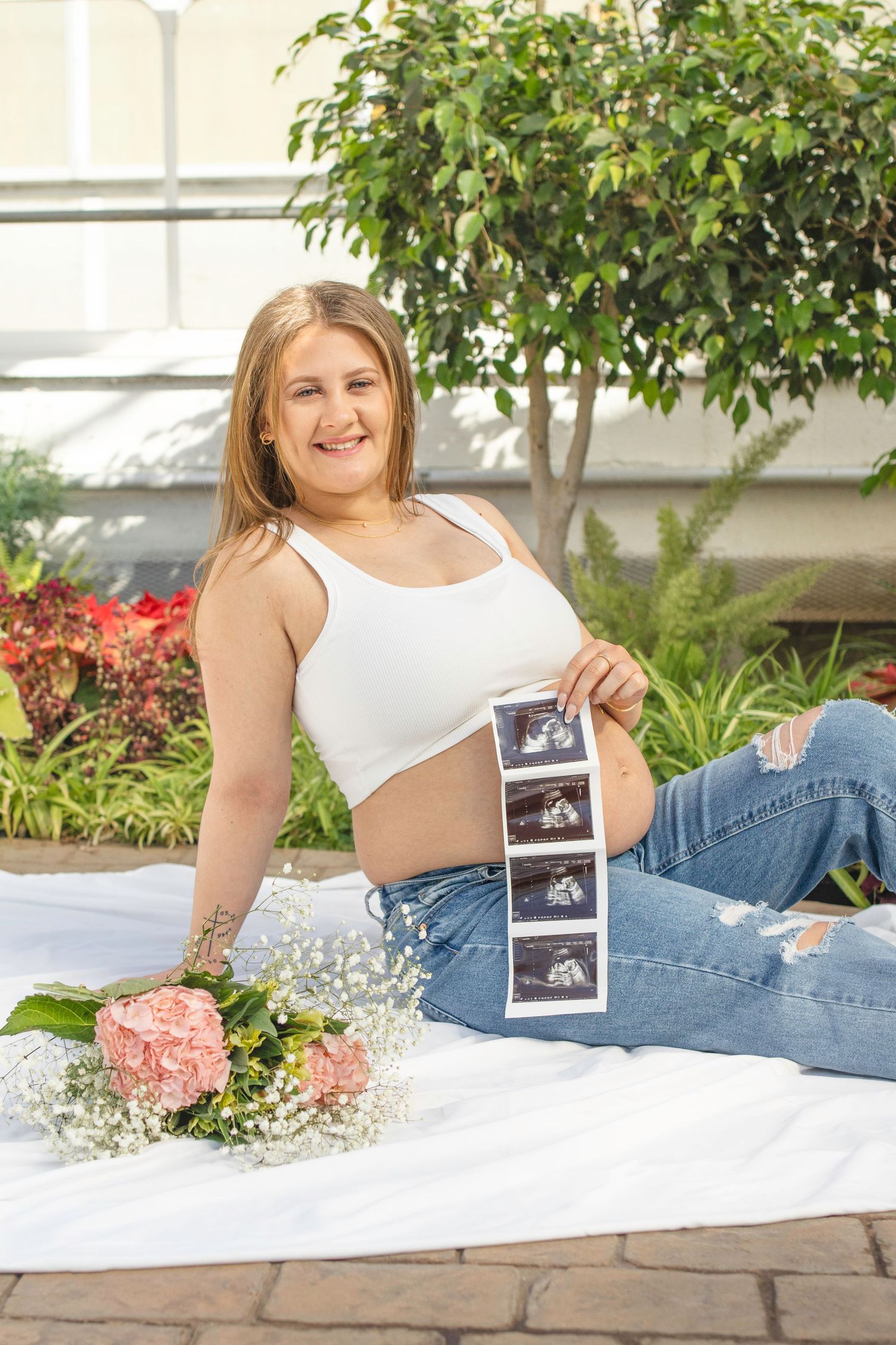 Maternity photo with ultrasound strips and flowers on white blanket in greenhouse setting.