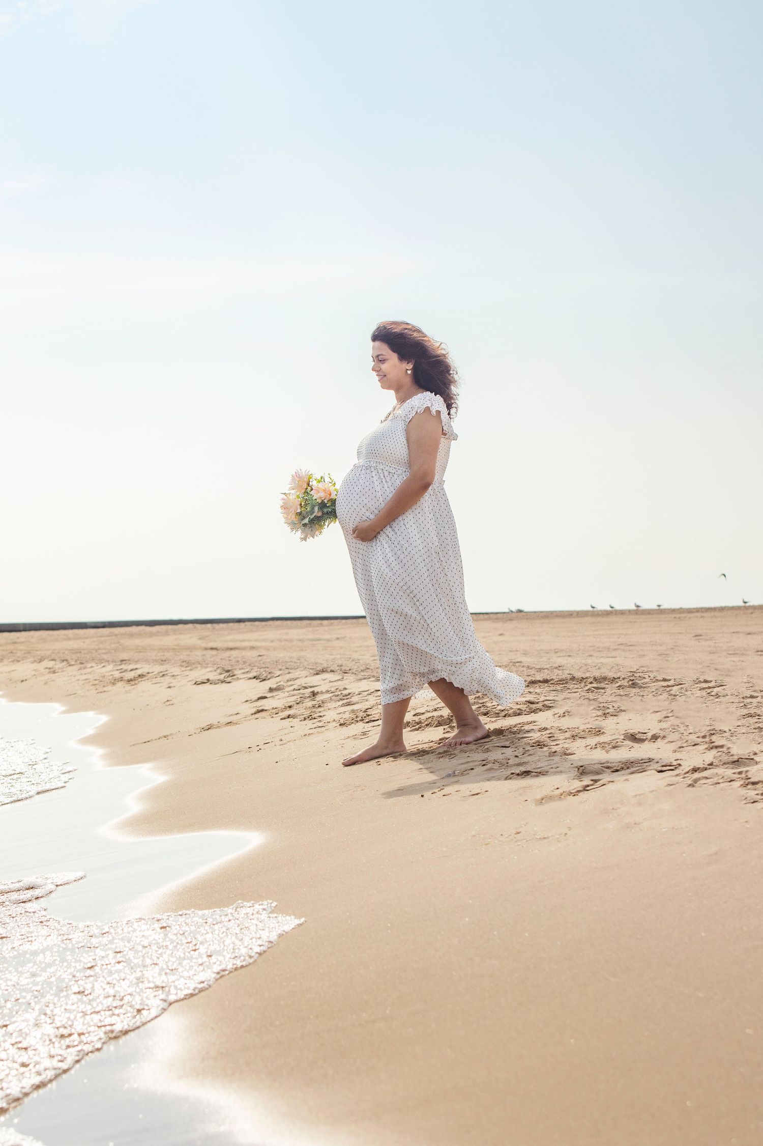A bride in a flowing white dress stands contemplatively at the ocean's edge on a serene beach at sunset.