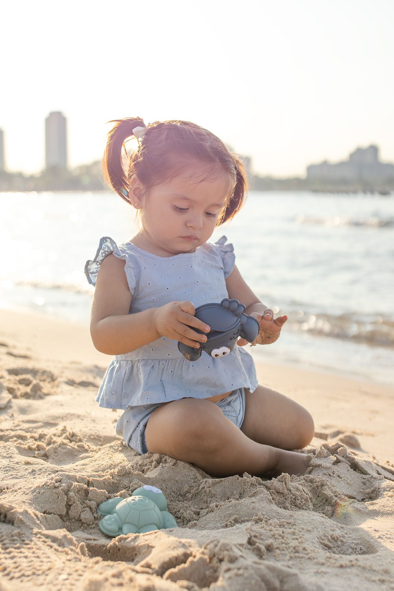 Cute toddler girl in blue dress plays with toy turtle in sand on sunny beach near water.