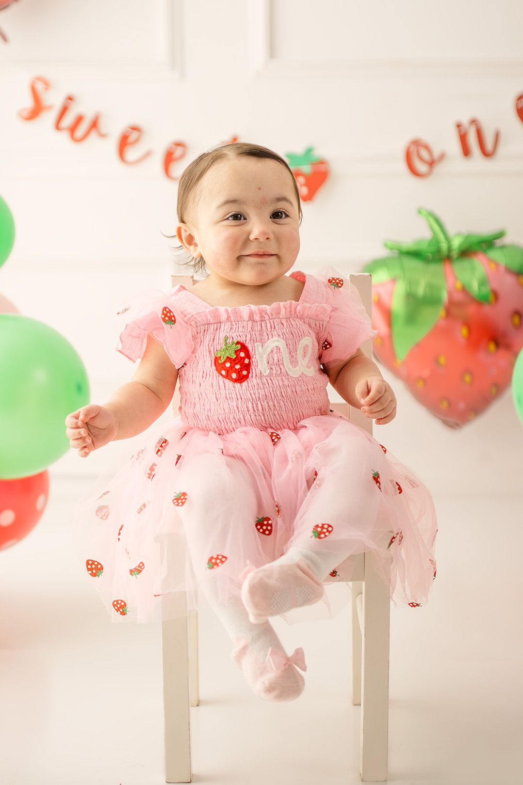 Baby girl in pink strawberry dress celebrates first birthday with balloons and Sweet One banner.