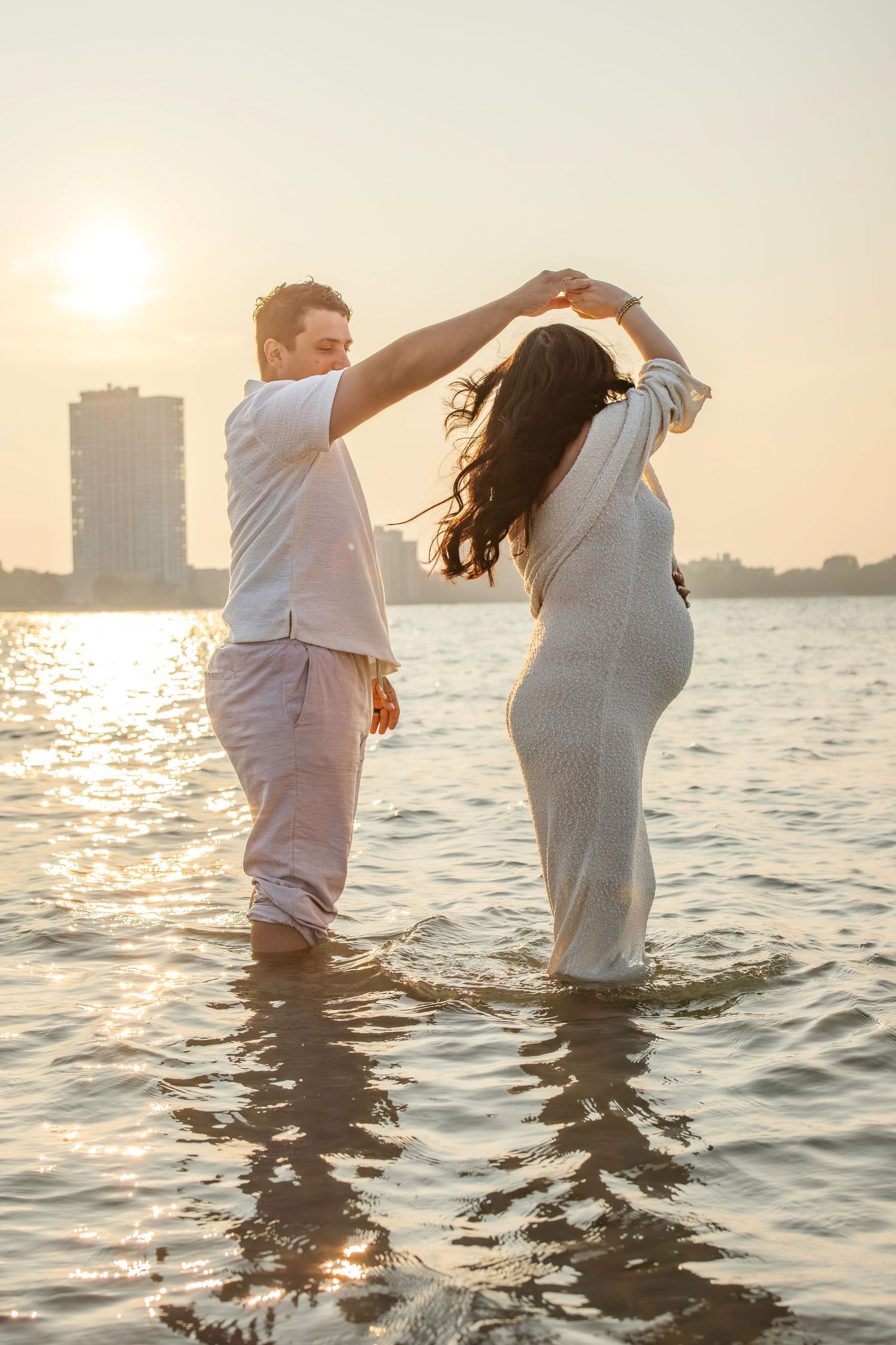 Couple in white outfits dancing in shallow water at sunset with city skyline in background.
