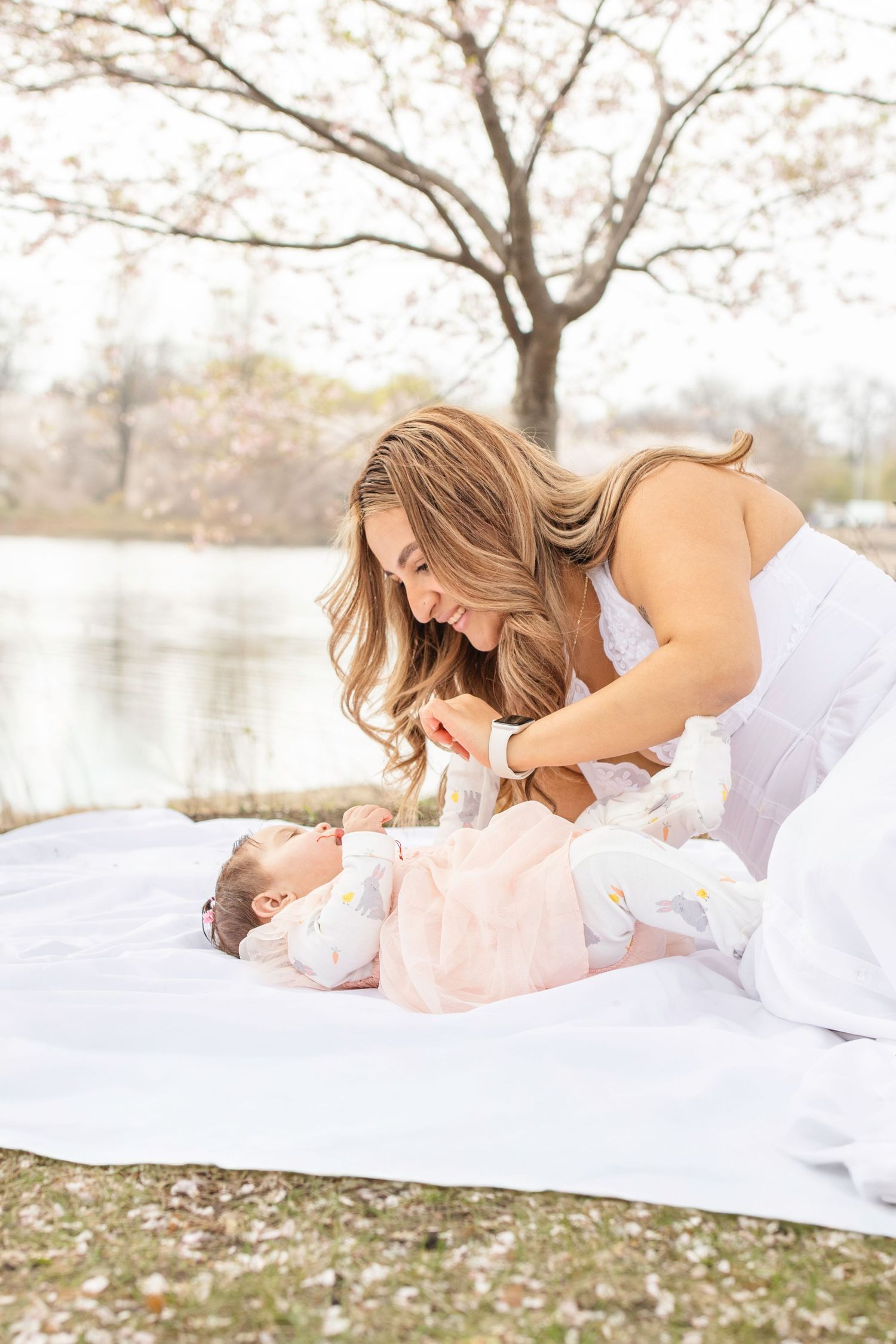 A tender spring outdoor moment between parent and baby on a white blanket under blooming cherry trees by a lake.