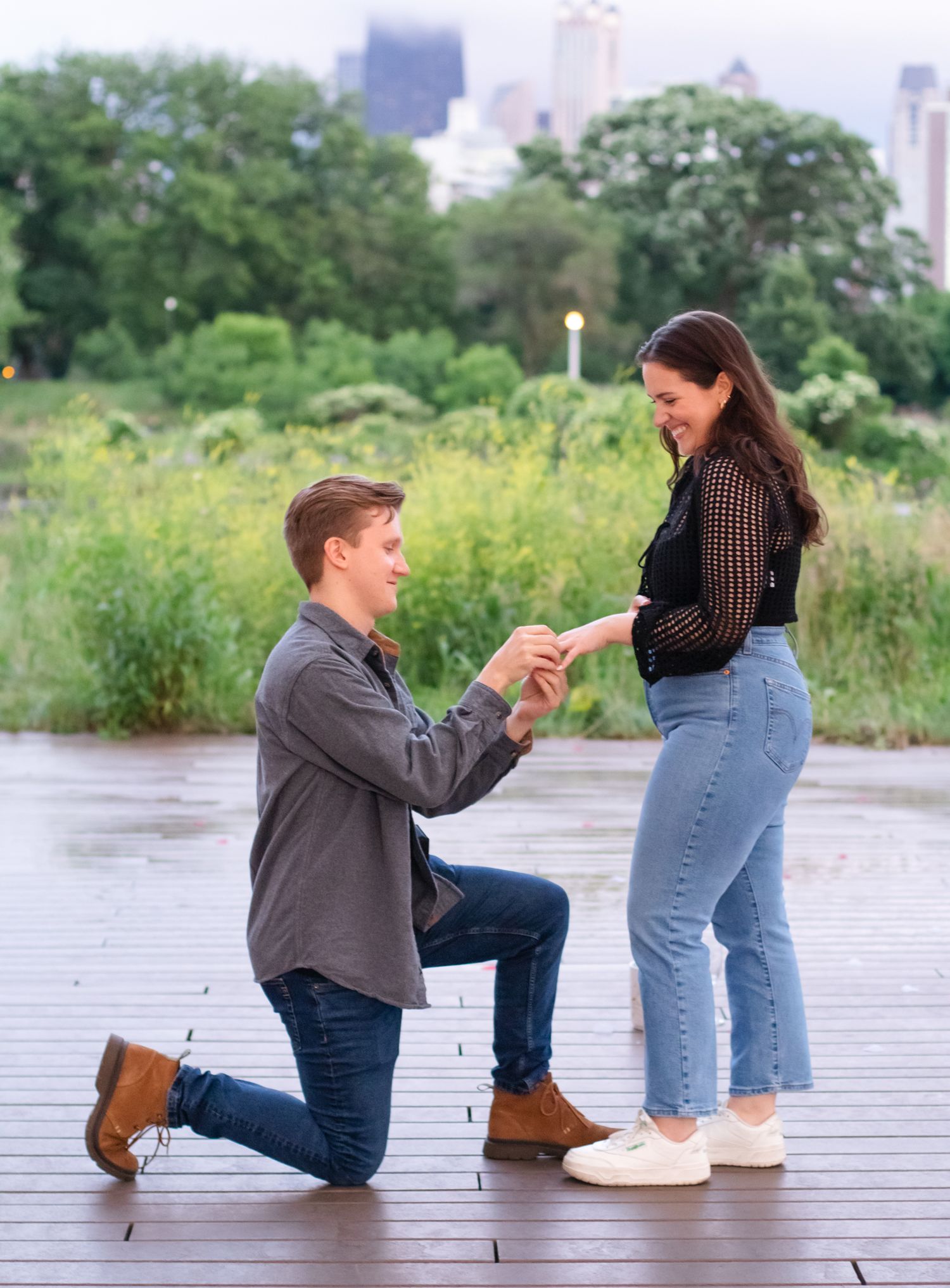 Surprise proposal moment captured as person kneels on wooden deck with city skyline backdrop.