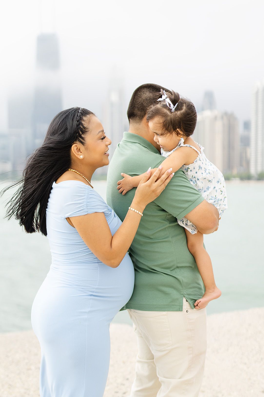 Pregnant woman in blue dress with husband holding toddler daughter by Chicago lakefront skyline.