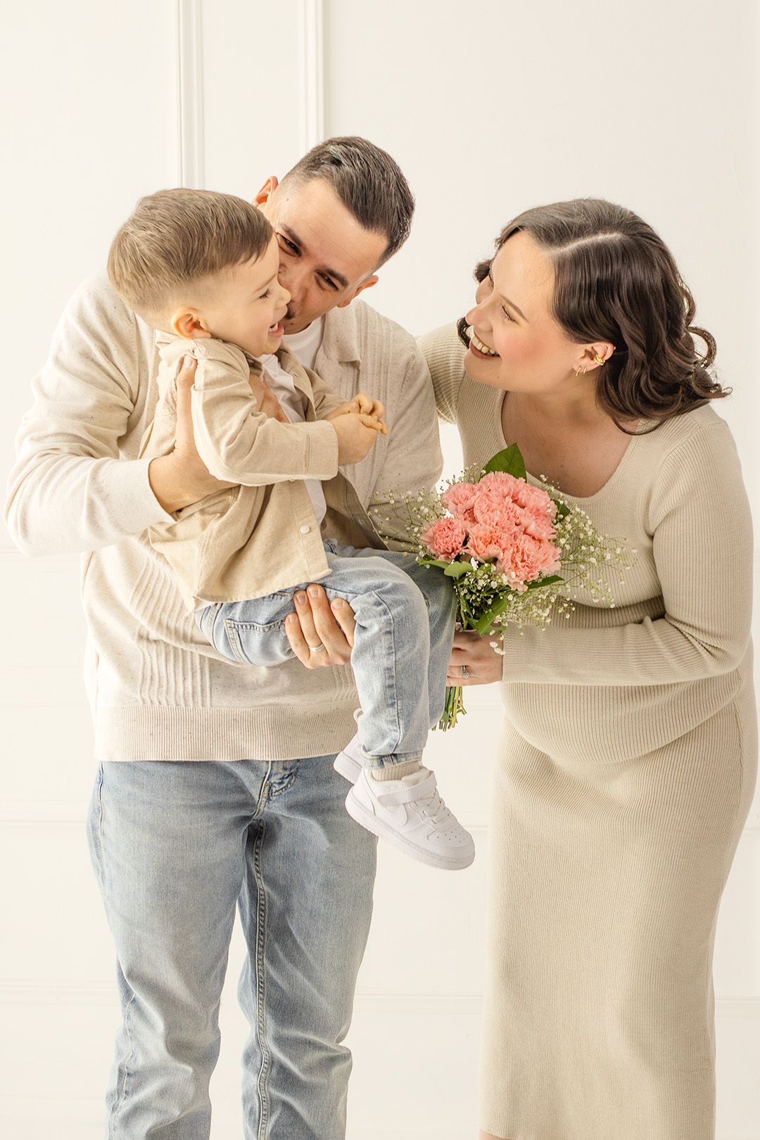 Smiling father holds toddler boy as pregnant mother laughs holding pink roses bouquet in white room.
