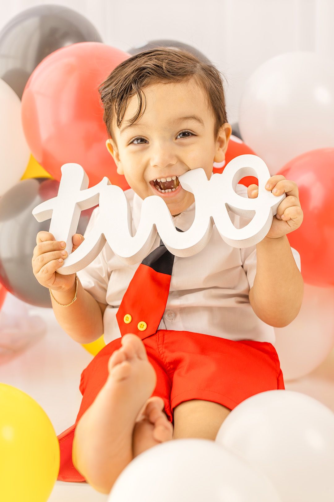 Happy toddler boy holds white 'two' sign laughing among colorful balloons at Mickey Mouse birthday party.