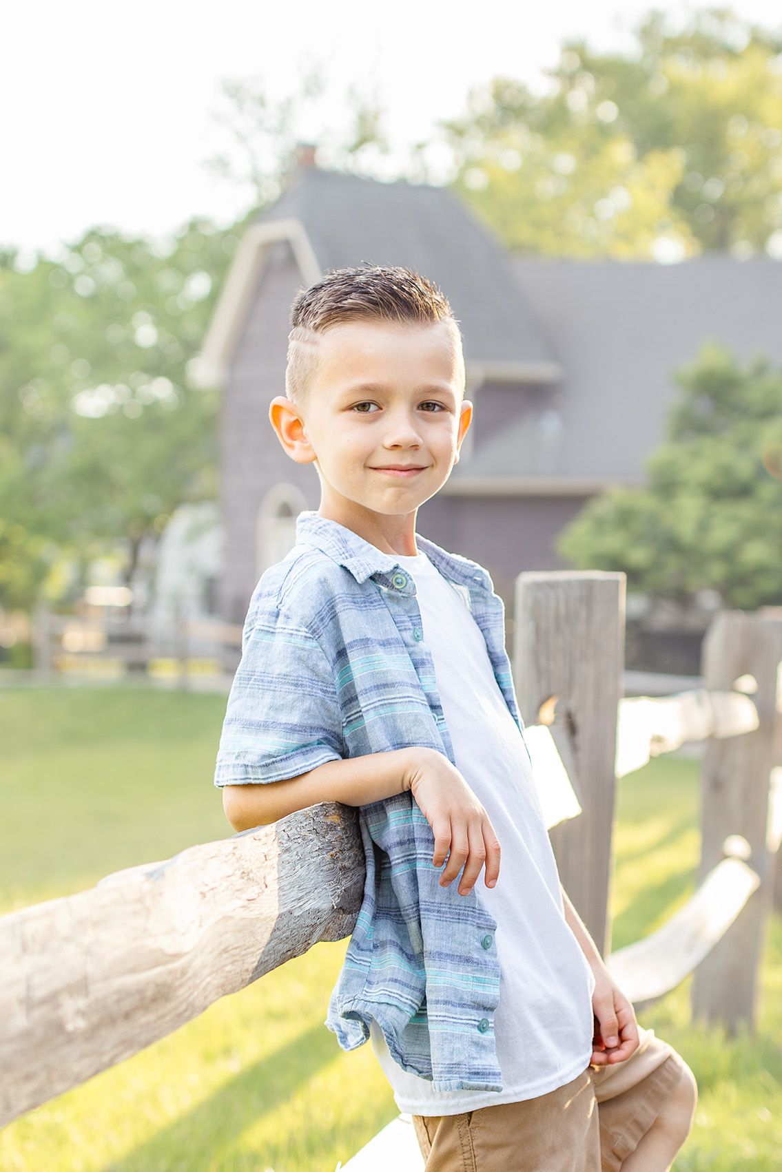 A young boy in a plaid shirt leans on a wooden fence, smiling outdoors near a house.