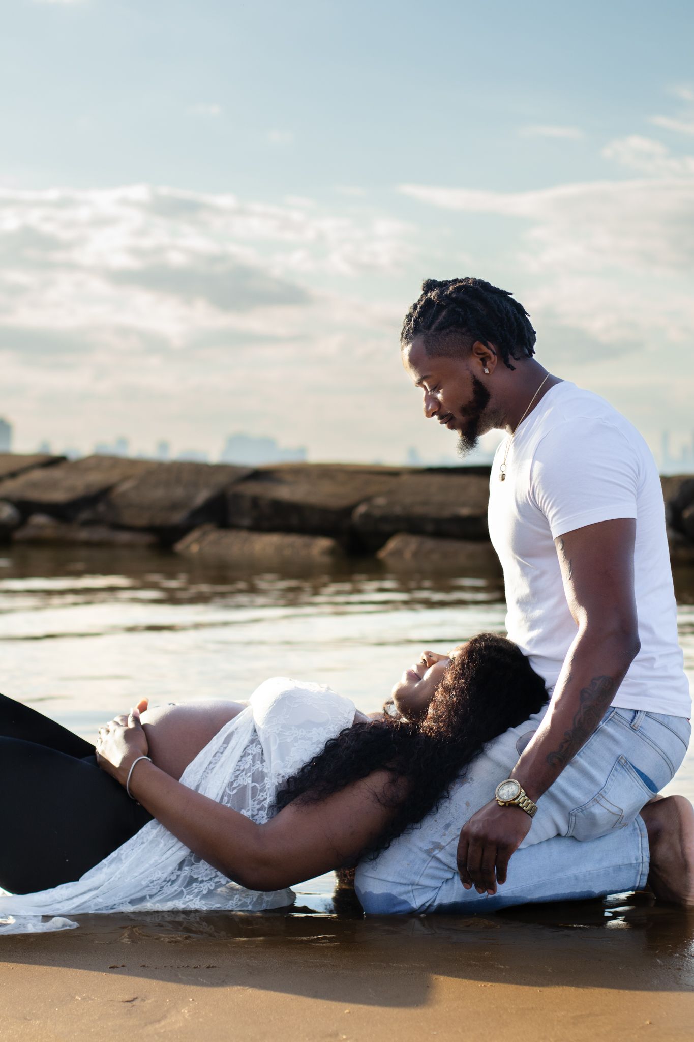 Romantic sunset maternity photoshoot on a beach with couple in white outfits lying on the sand near water.
