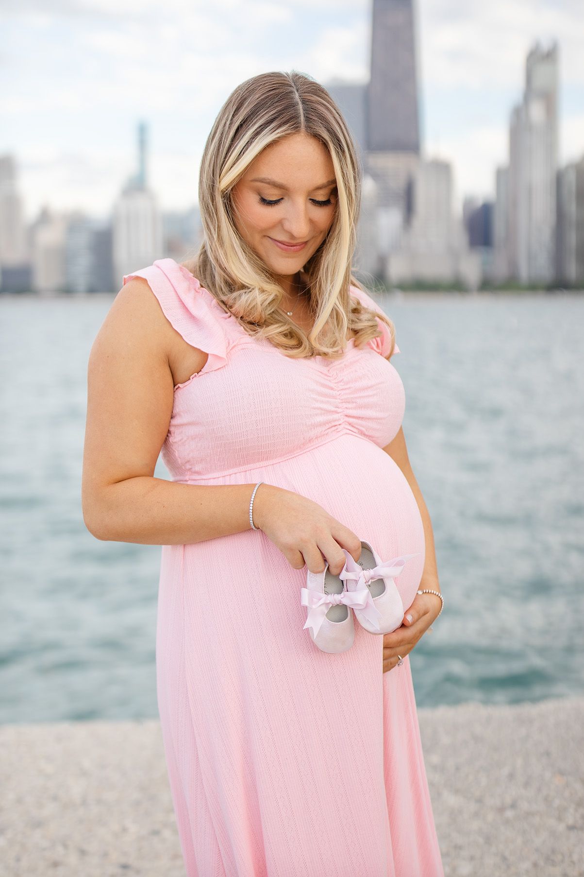 Pregnant woman in pink dress holding baby shoes by Chicago skyline waterfront.