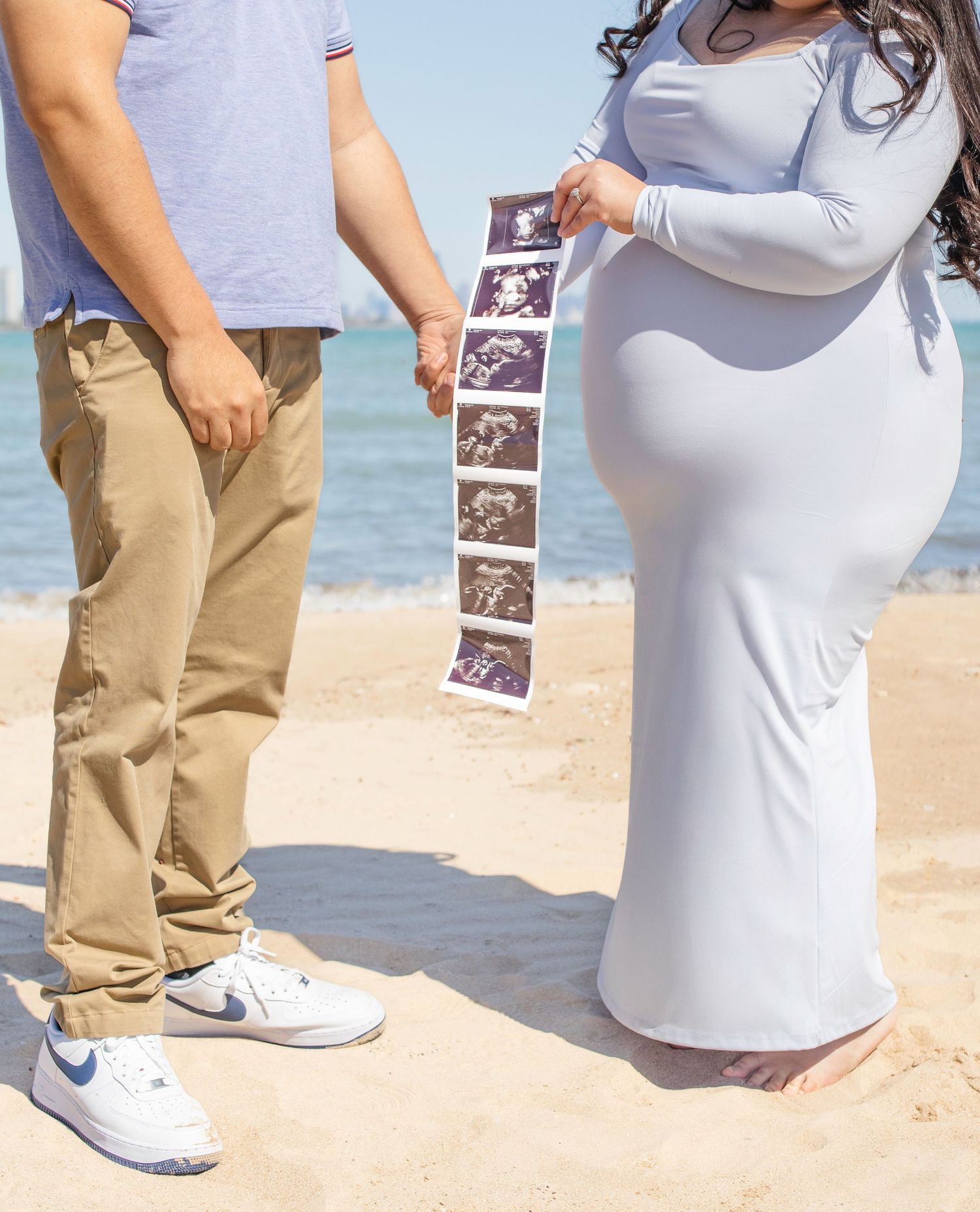 A couple on the beach holds a strip of ultrasound photos while standing by the water's edge.