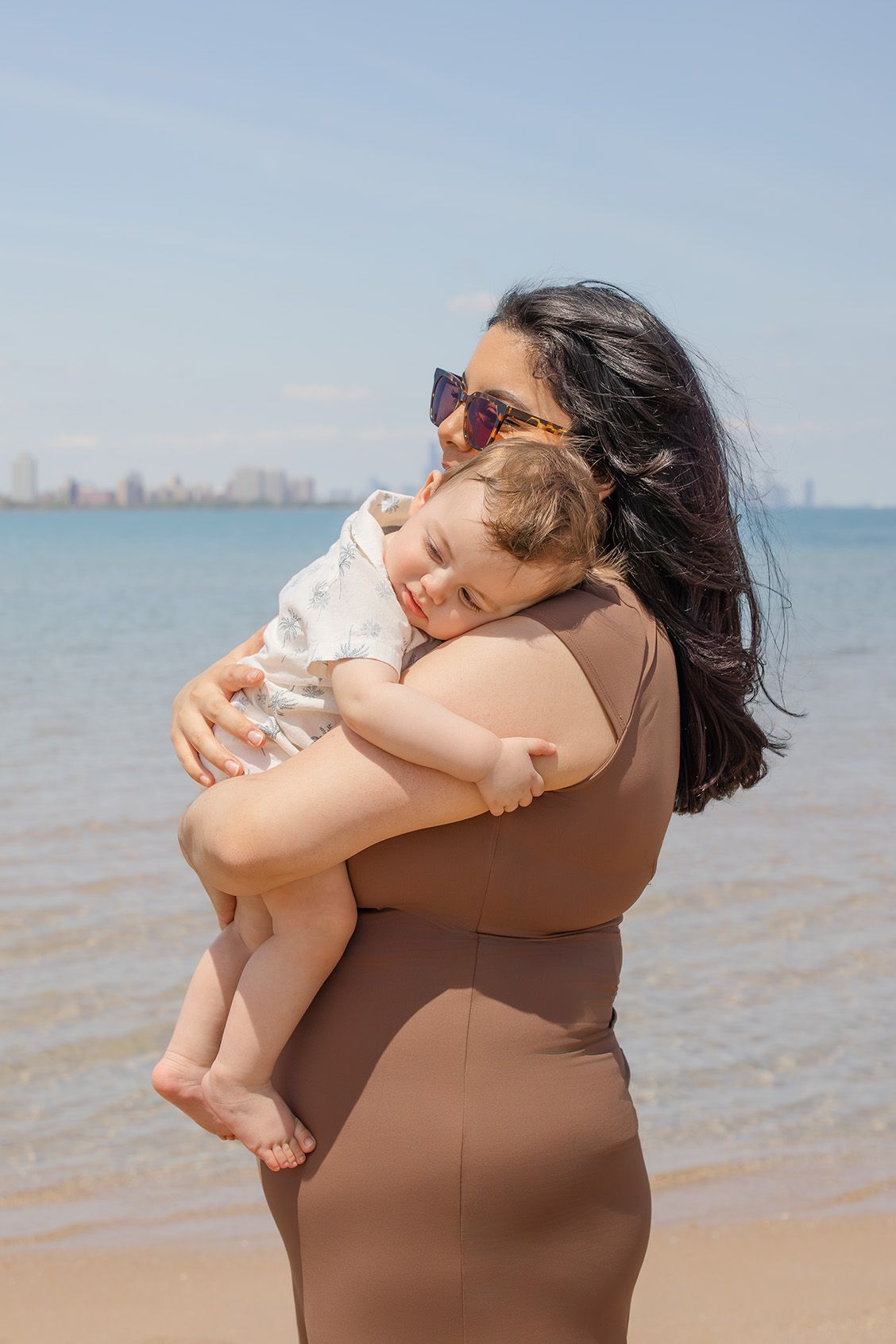 A woman in a brown swimsuit holds a sleeping baby on a sunny beach with blue water in the background.