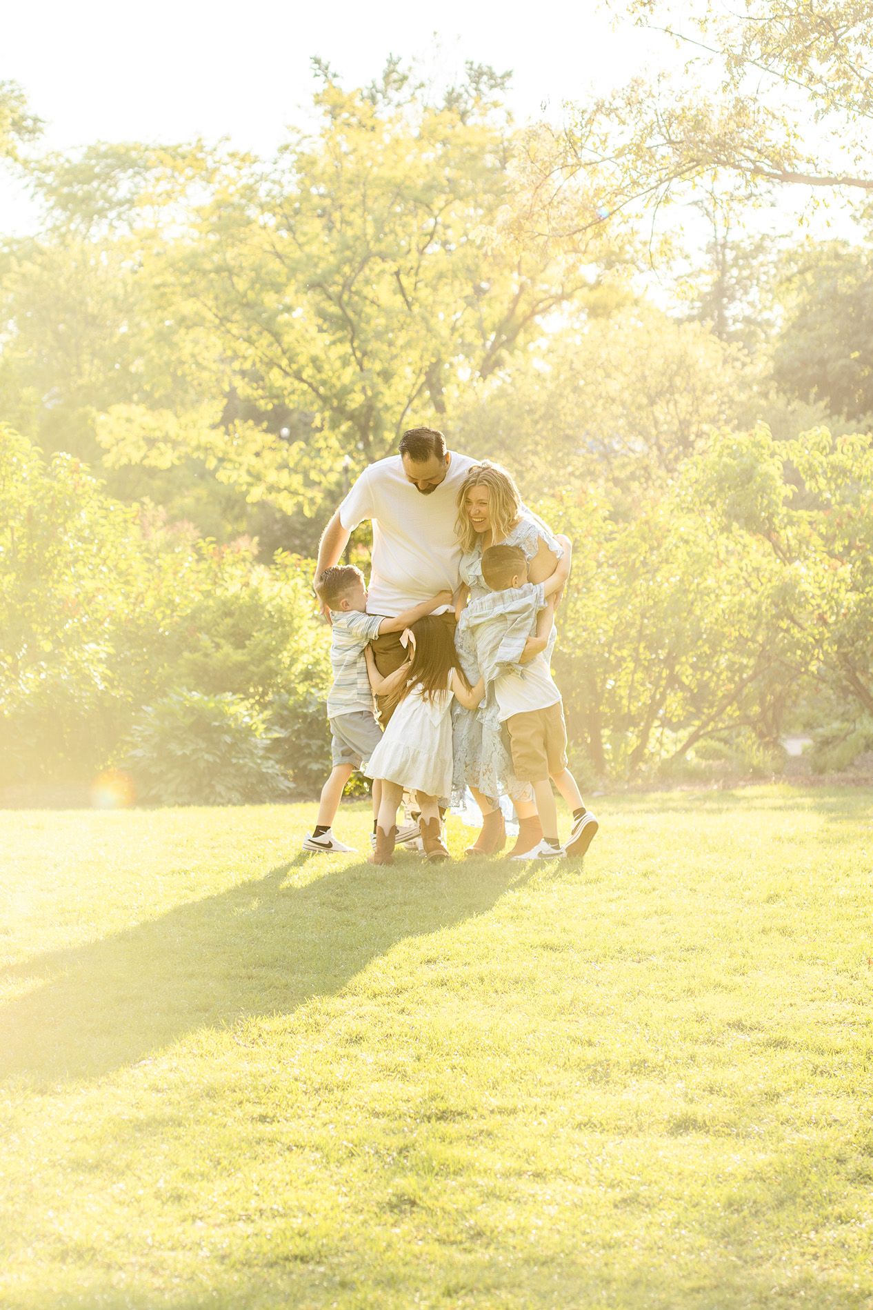 Family hugging and playing together on green grass in a sunlit park surrounded by lush trees.