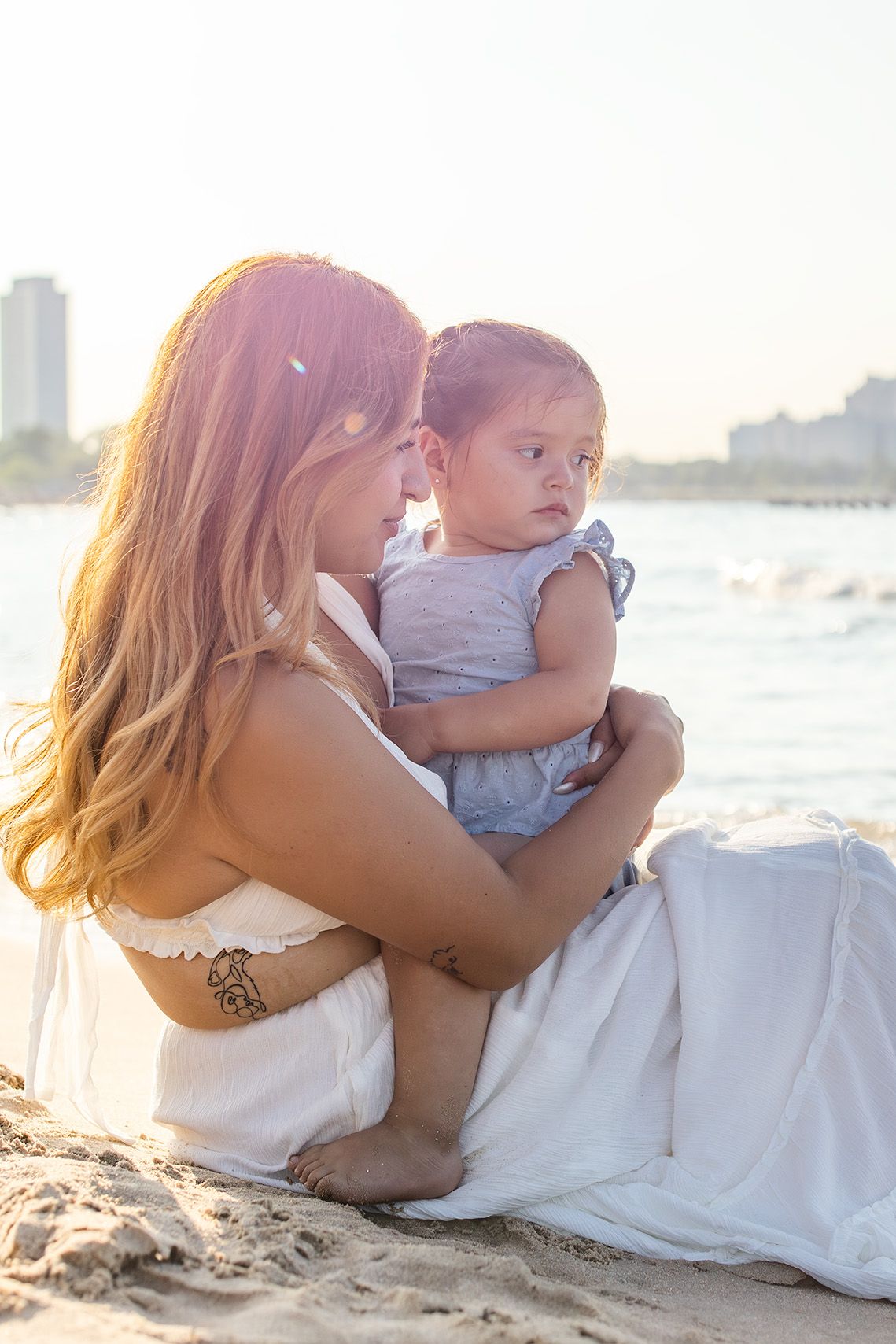 Mother sitting on sandy beach holding infant in blue outfit with golden sunset backlight.