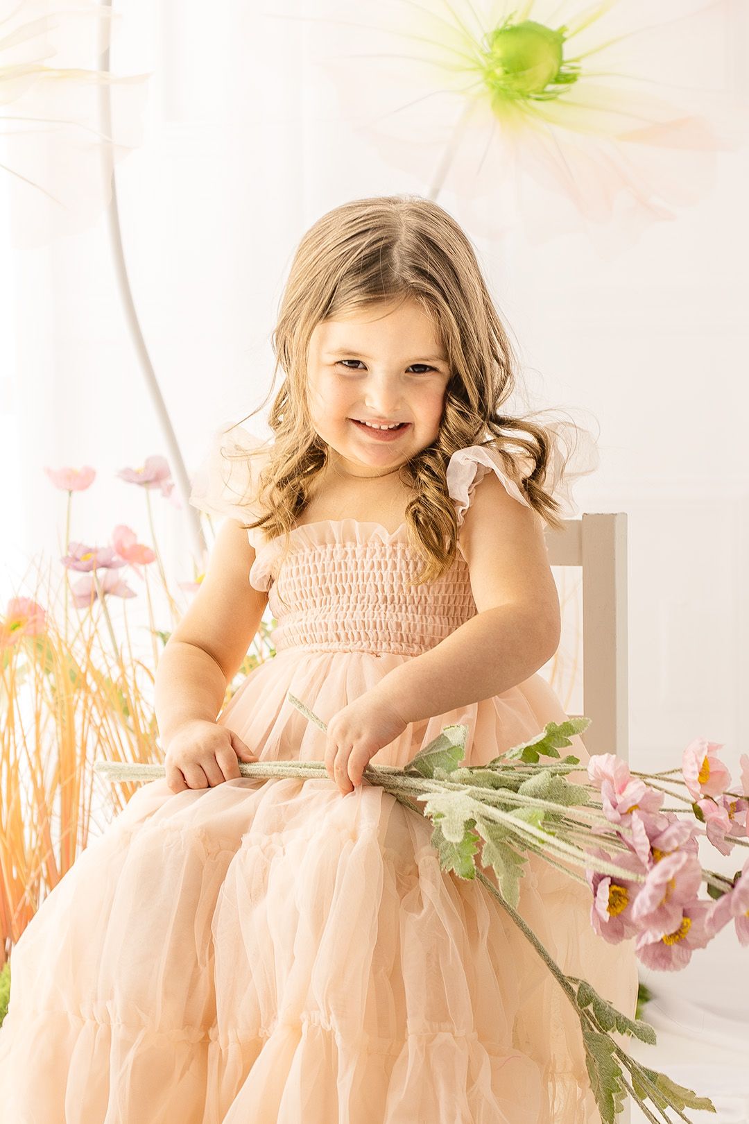A child in a peach tulle dress holds pink flowers while sitting on a white chair in a dreamy spring setting.