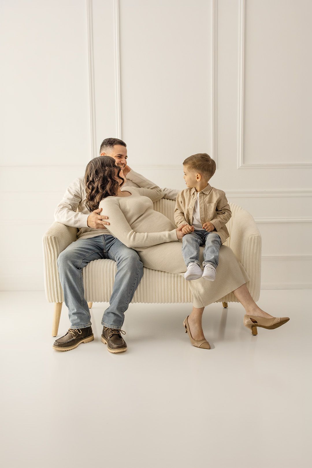 A happy family of three sitting on a beige sofa, with a pregnant mother, father, and young son in a bright white room.