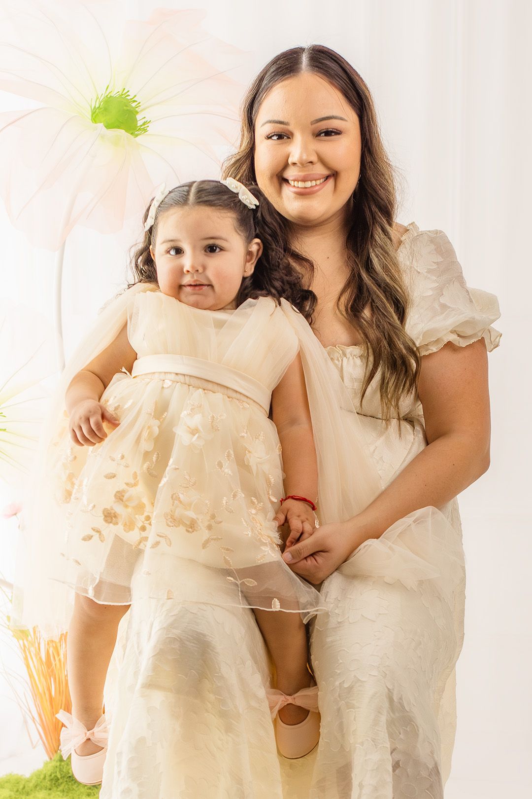 A smiling young woman in a cream dress holds a toddler girl in a matching outfit against a floral backdrop.
