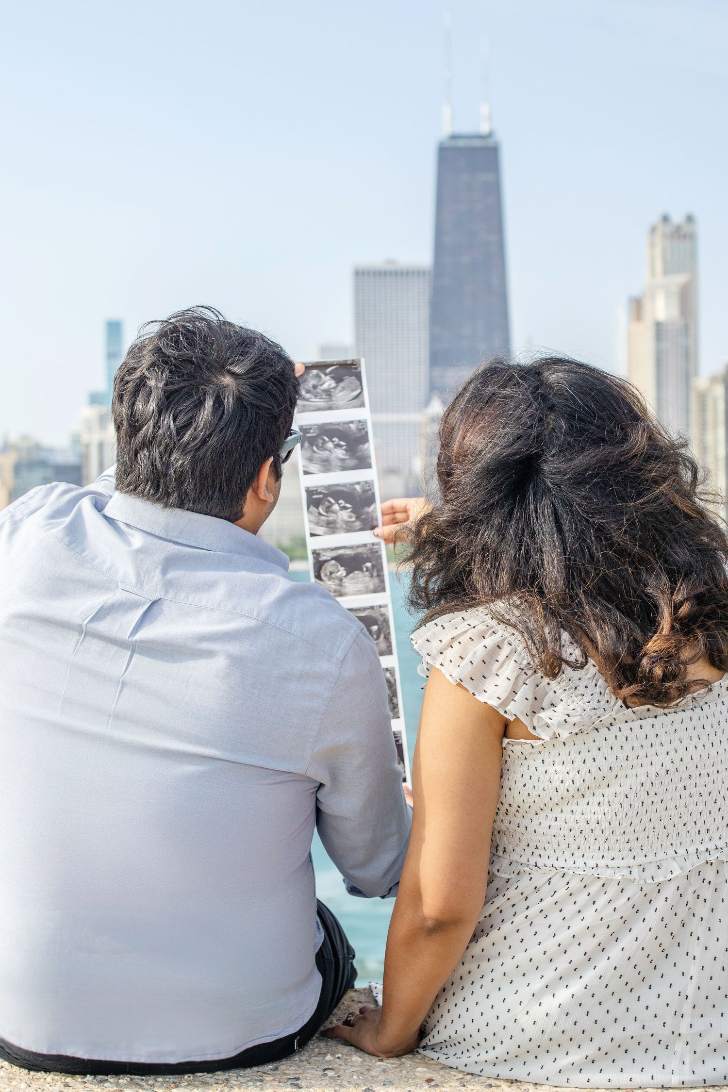 A couple seated on a rooftop holds ultrasound photos while looking at the Chicago skyline on a sunny day.