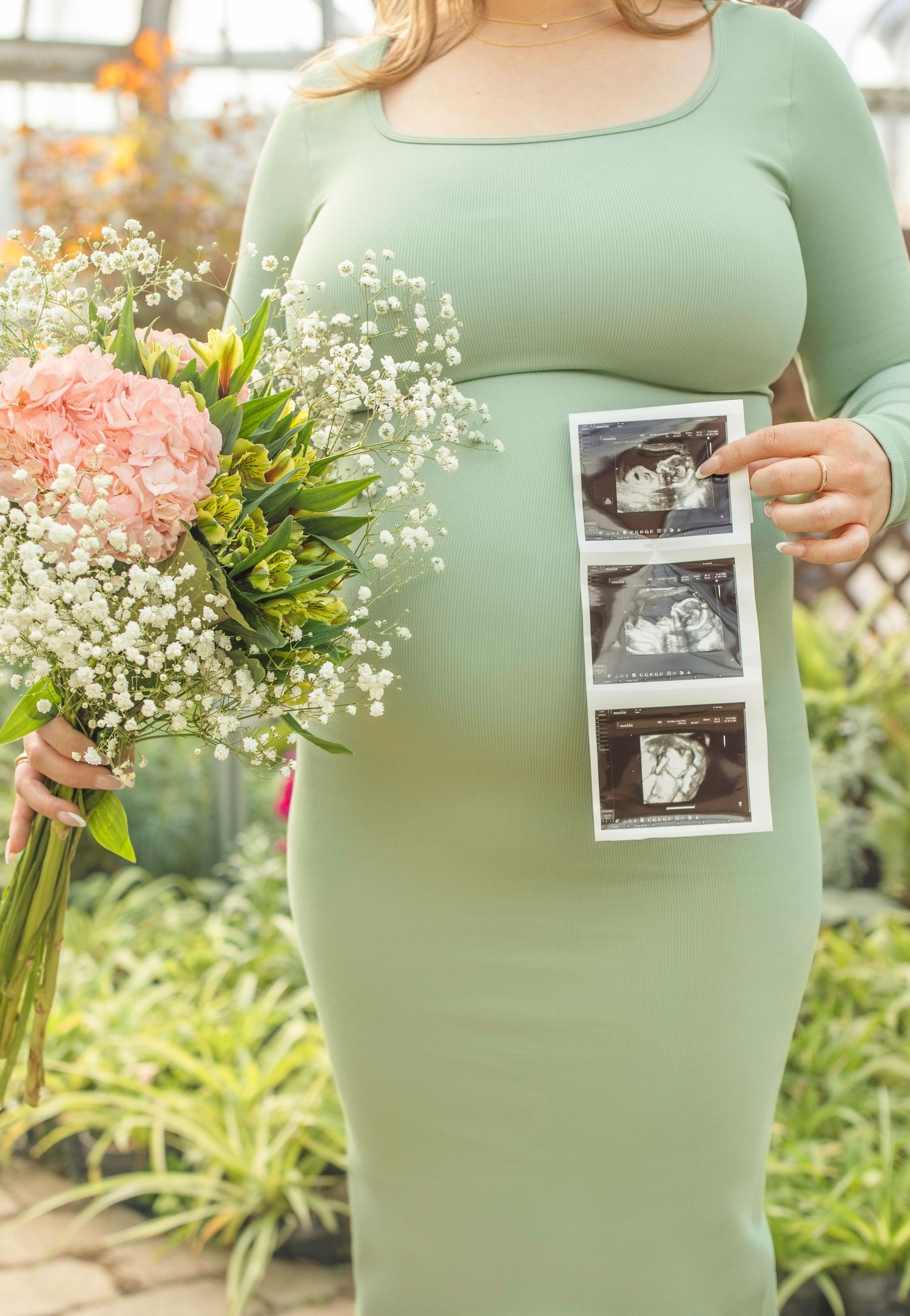Pregnant woman in green dress holds ultrasound photos and floral bouquet in greenhouse setting.