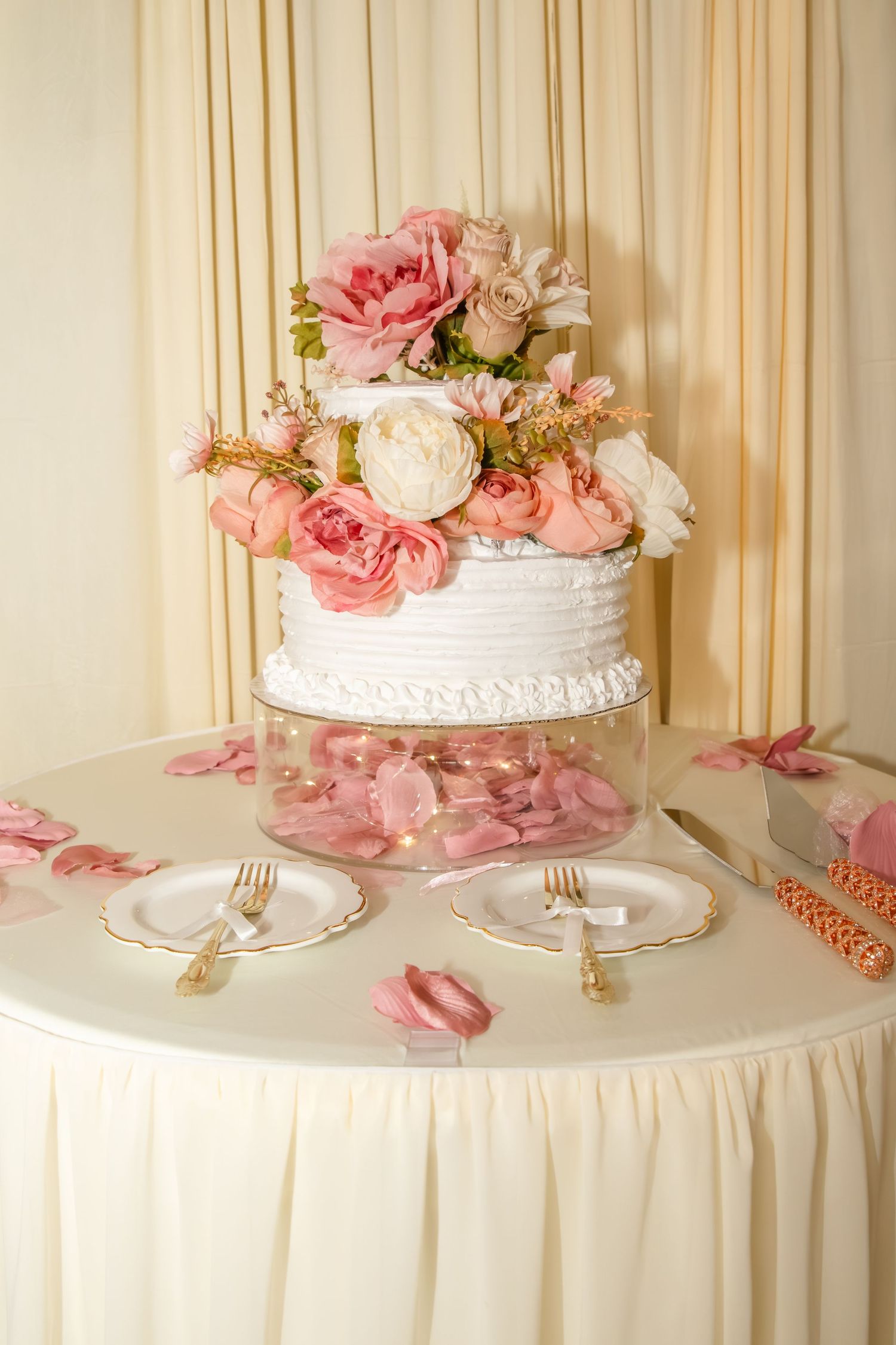 Elegant two-tier white wedding cake adorned with pink roses and peonies on a draped table with gold forks and rose petals.