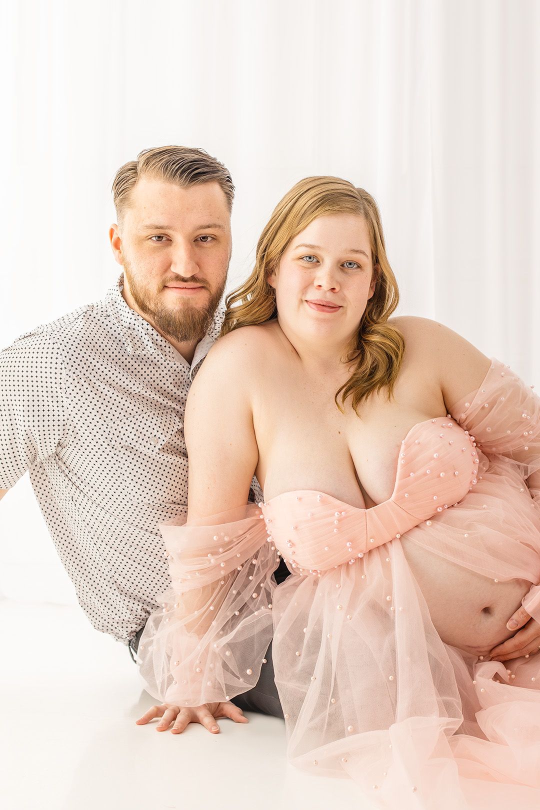 Couple poses together during maternity photoshoot with woman wearing pink tulle gown.