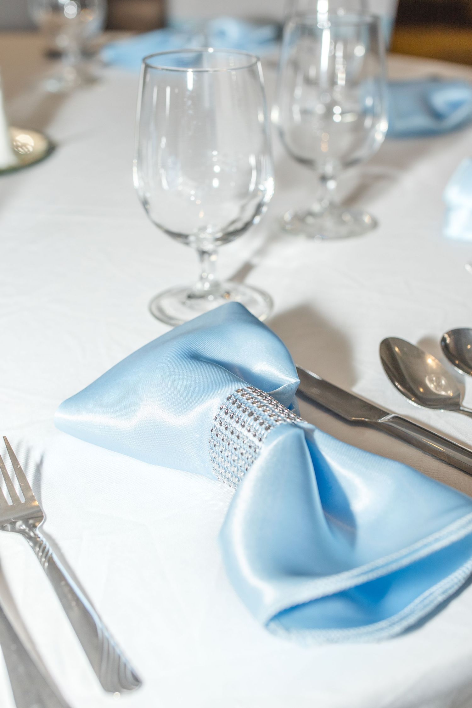 An elegant table setting with light blue napkin, crystal stemware and rhinestone napkin ring on white tablecloth.