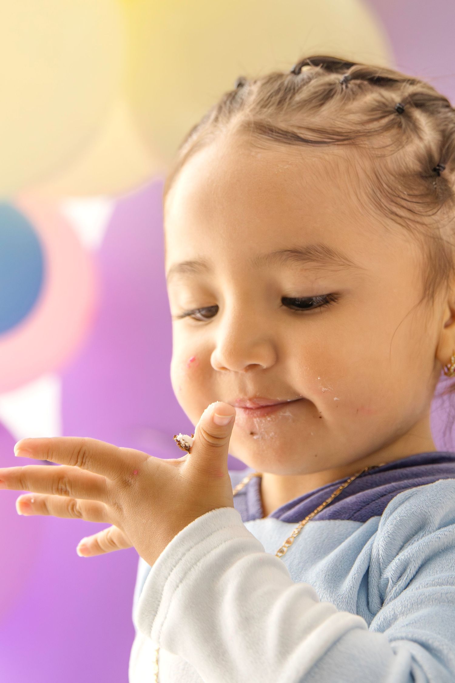 A young girl with braids smiles while licking frosting off her finger at a colorful birthday party.