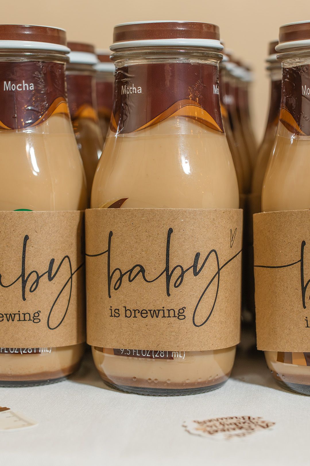 Baby shower mocha coffee bottles with kraft paper labels reading baby is brewing, arranged as party favors.