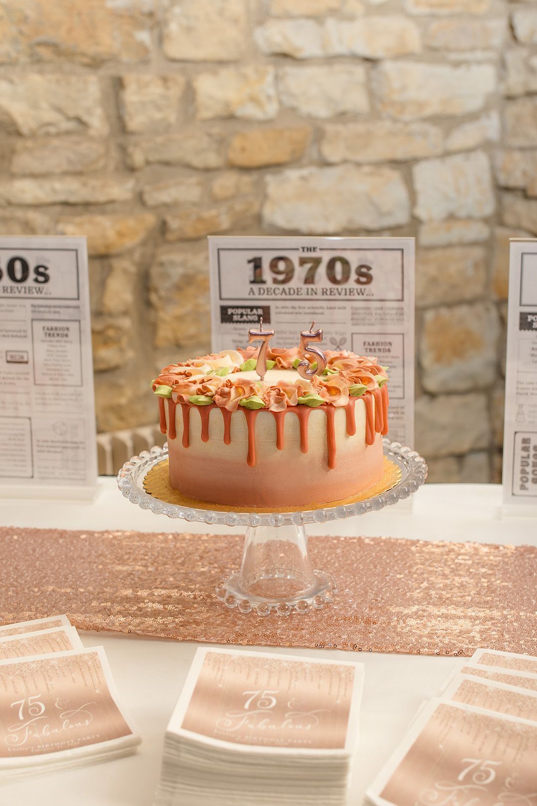 A 75th birthday cake on a pedestal stand with a 1970s decade review backdrop at a rose gold themed party.