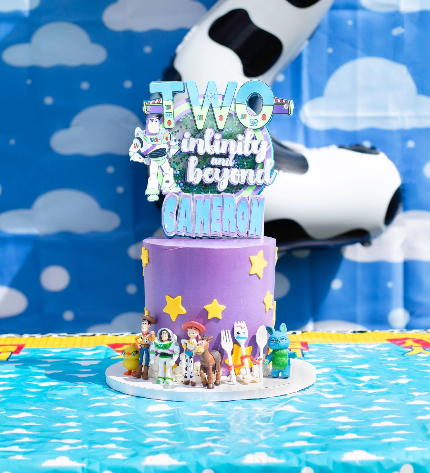Purple birthday cake with Toy Story figurines and custom topper against blue cloud backdrop.
