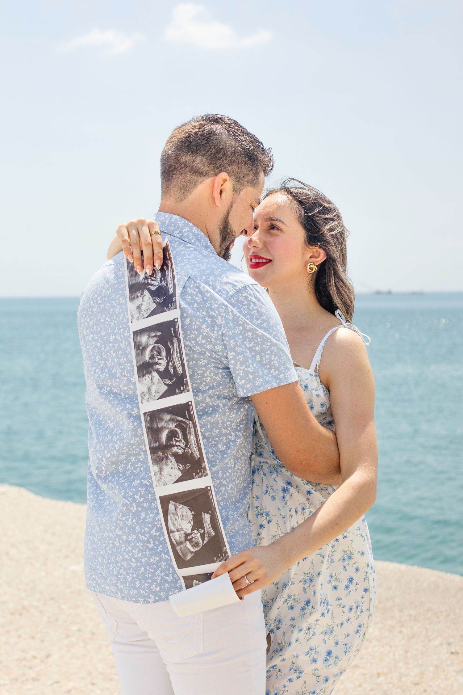 Couple celebrates pregnancy announcement on beach while holding ultrasound photos with turquoise ocean in background.