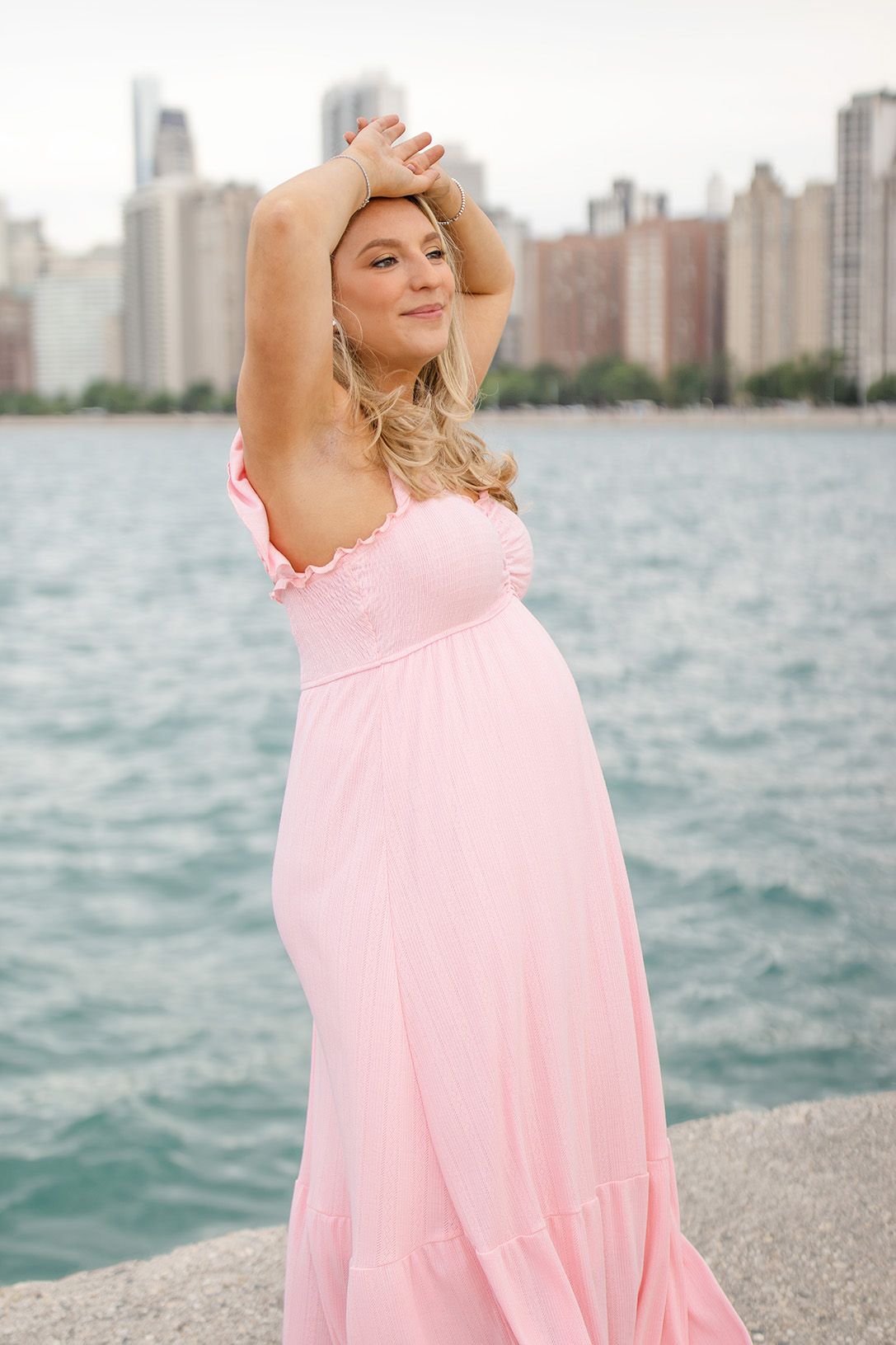 Pregnant woman in pink maxi dress posing by Lake Michigan with Chicago skyline in background.
