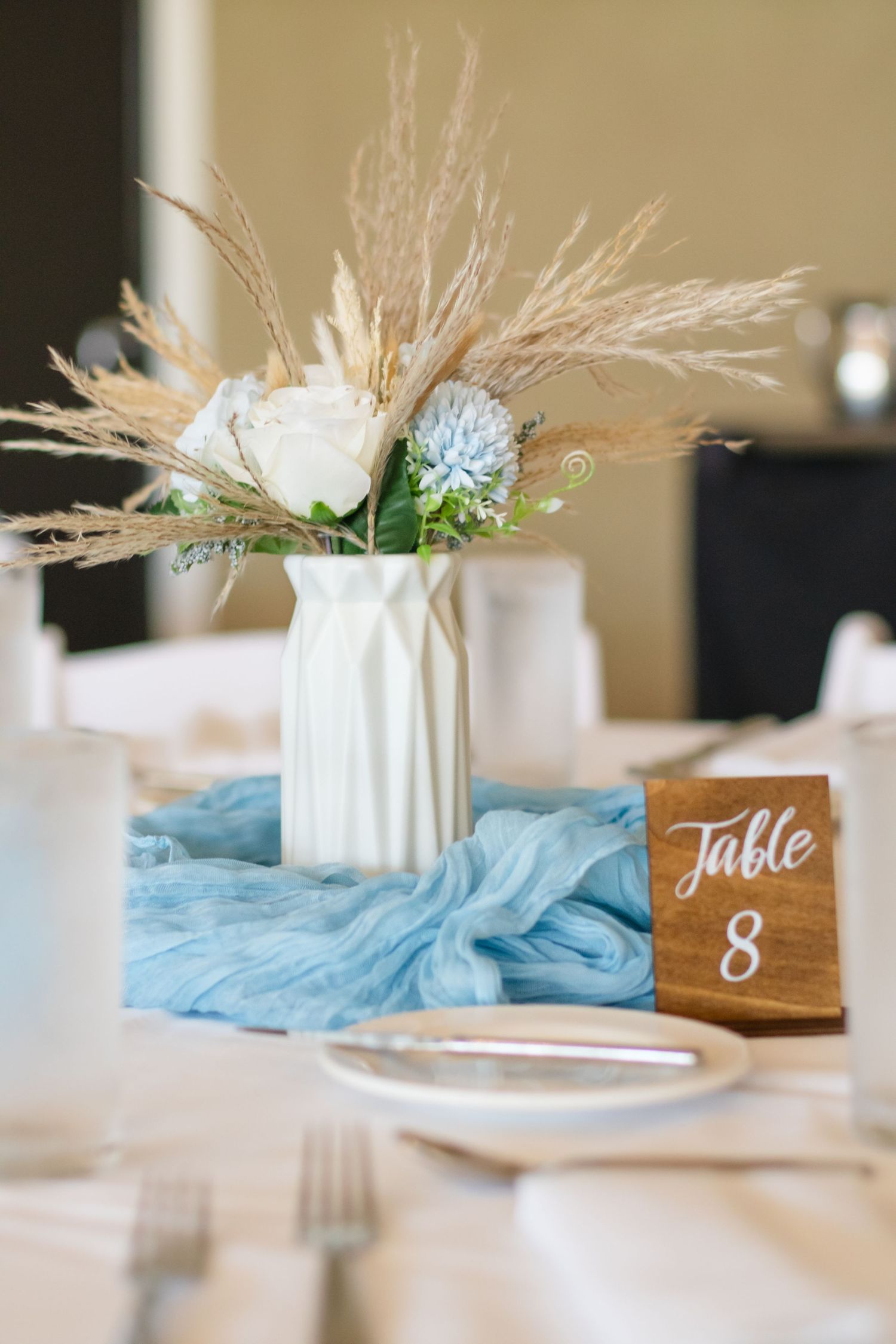 Wedding centerpiece with white vase, dried pampas grass, blue flowers and wooden table number 8 sign on light blue fabric.
