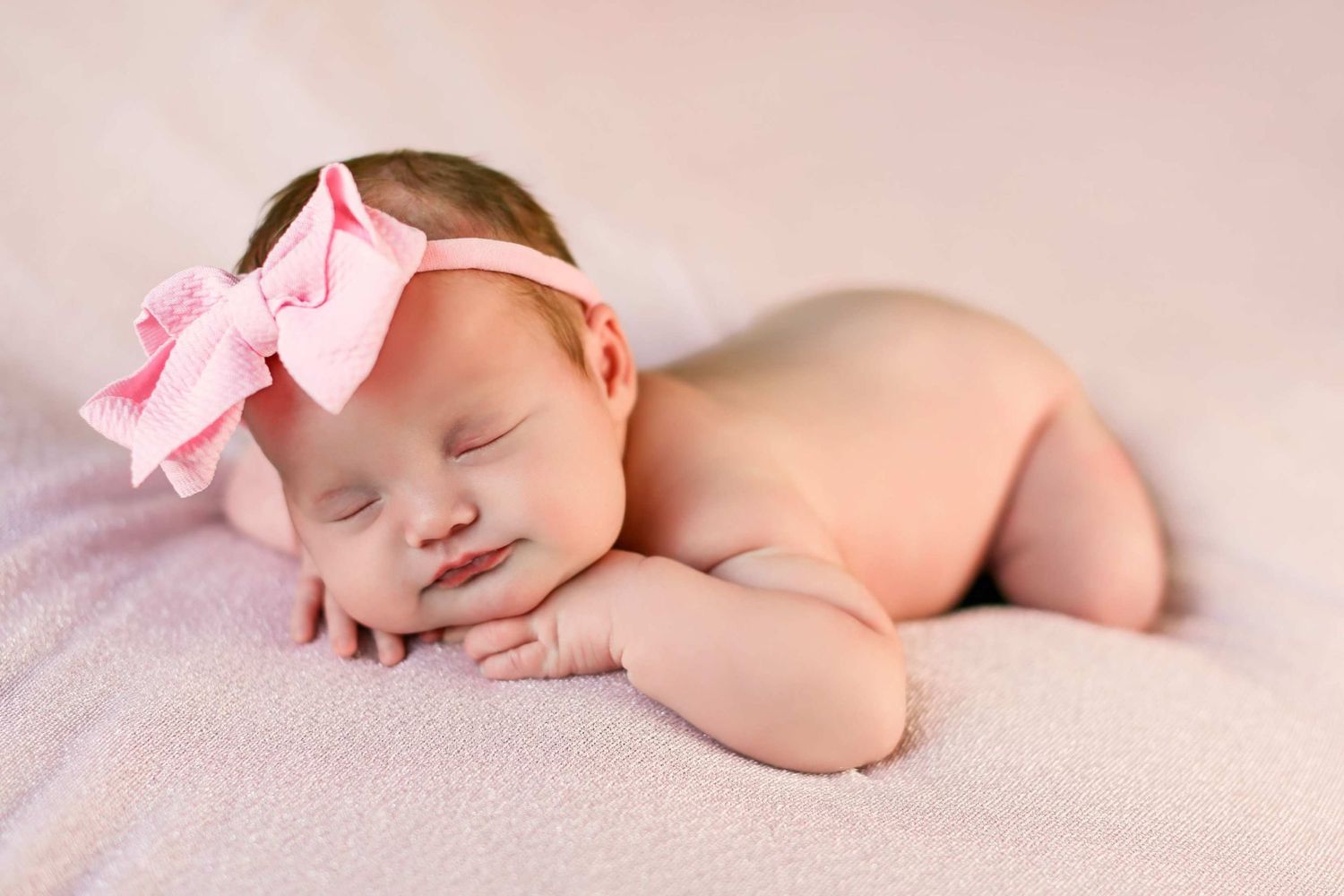 Sleeping newborn baby wearing pink bow headband lying on soft pink blanket in studio setting.