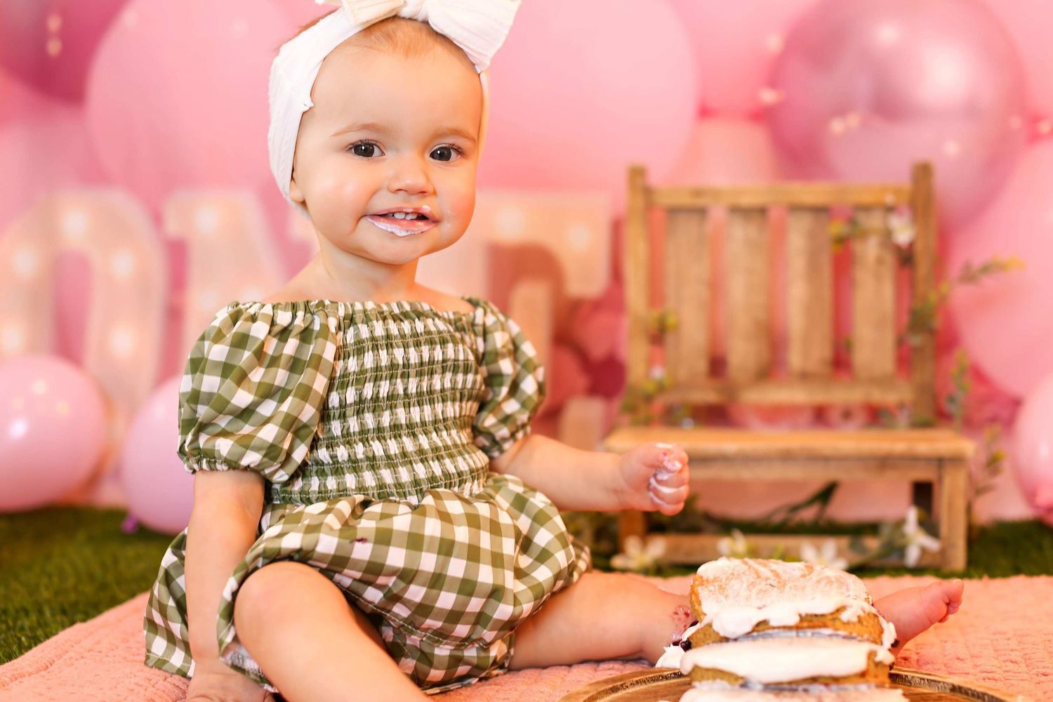 Baby in plaid outfit sits on pink backdrop during cake smash first birthday photo session with white frosted cake.