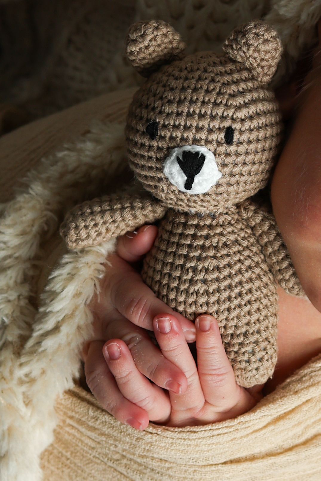 Newborn baby hands holding a crocheted teddy bear, wrapped in soft cream knit blanket.
