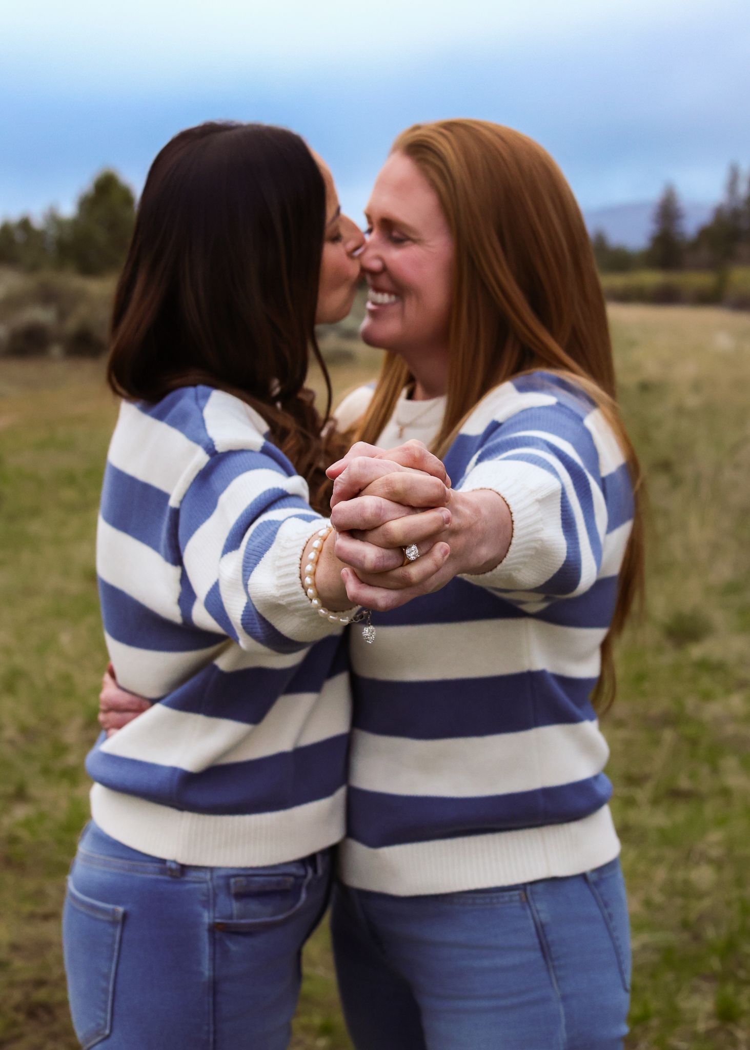 Two women in matching blue striped sweaters embrace and share a kiss outdoors in a field.