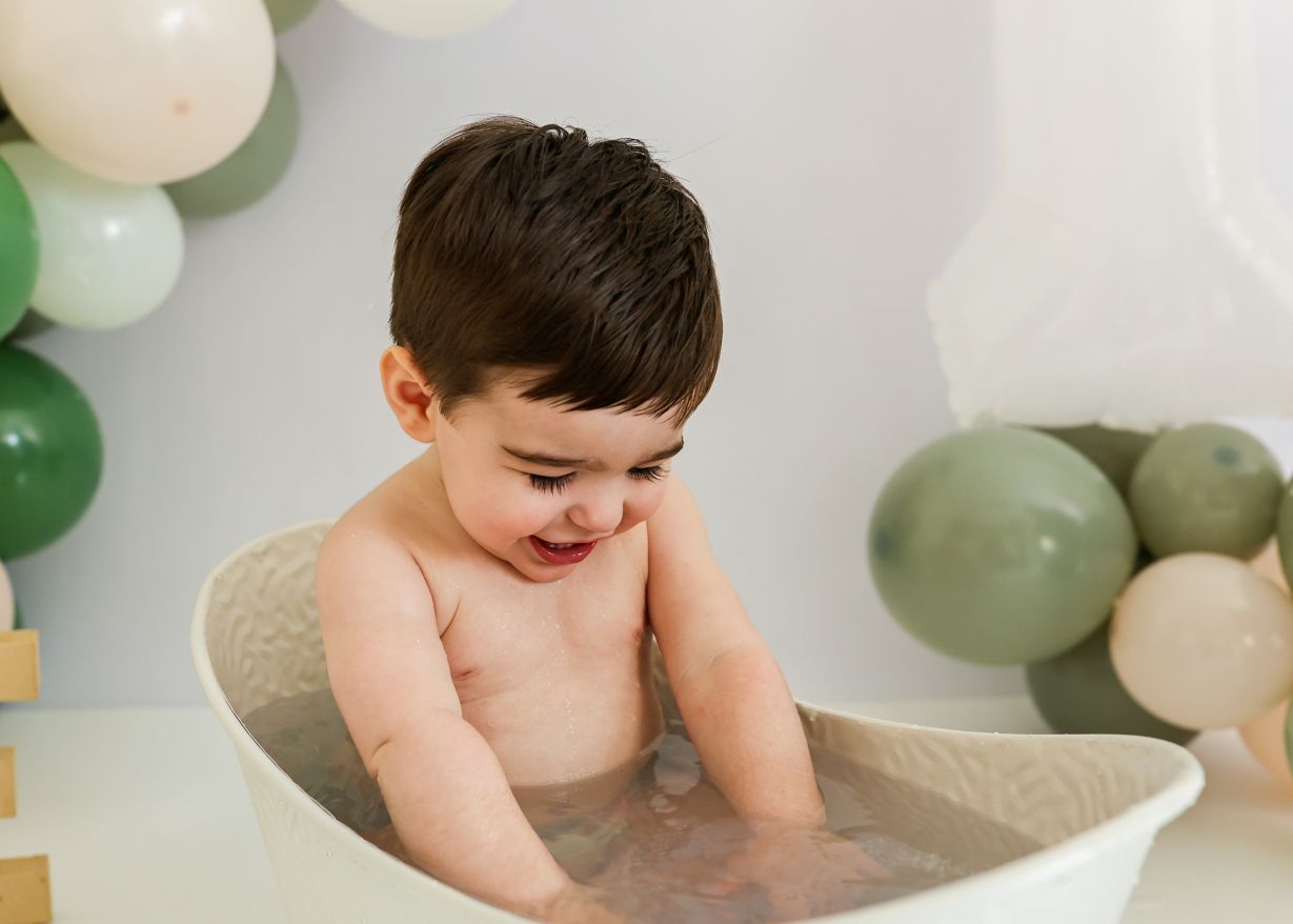 Smiling toddler boy with dark hair enjoys a bubble bath with green balloon backdrop.