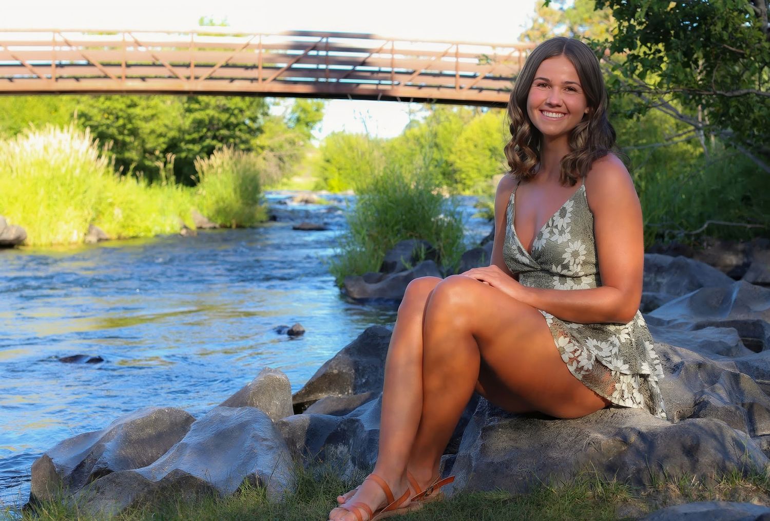 Young woman smiling while sitting on rocks beside a scenic creek with a wooden bridge in the background.