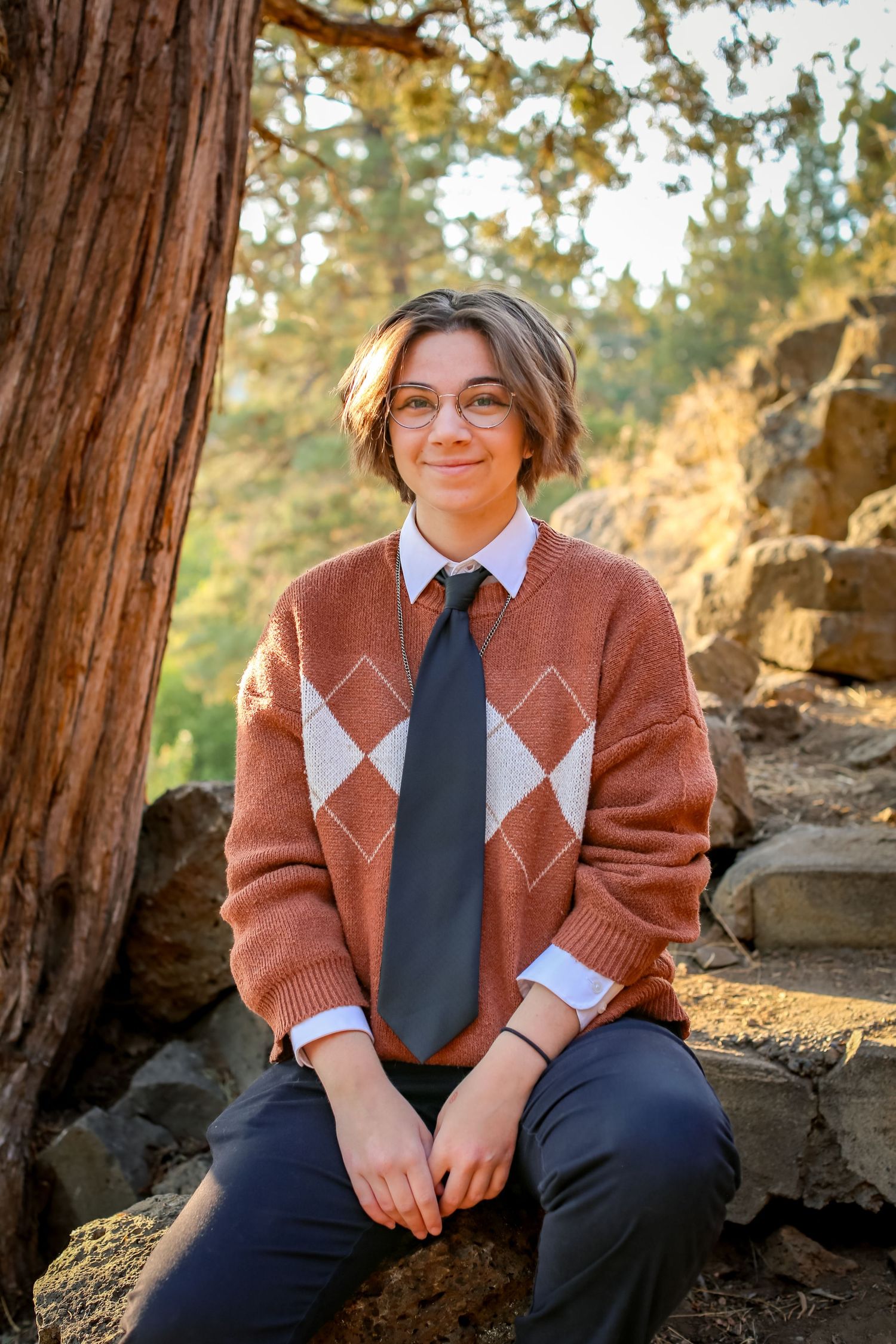 Teen in argyle sweater and tie sits on rocks outdoors, smiling, surrounded by trees and natural stone backdrop.
