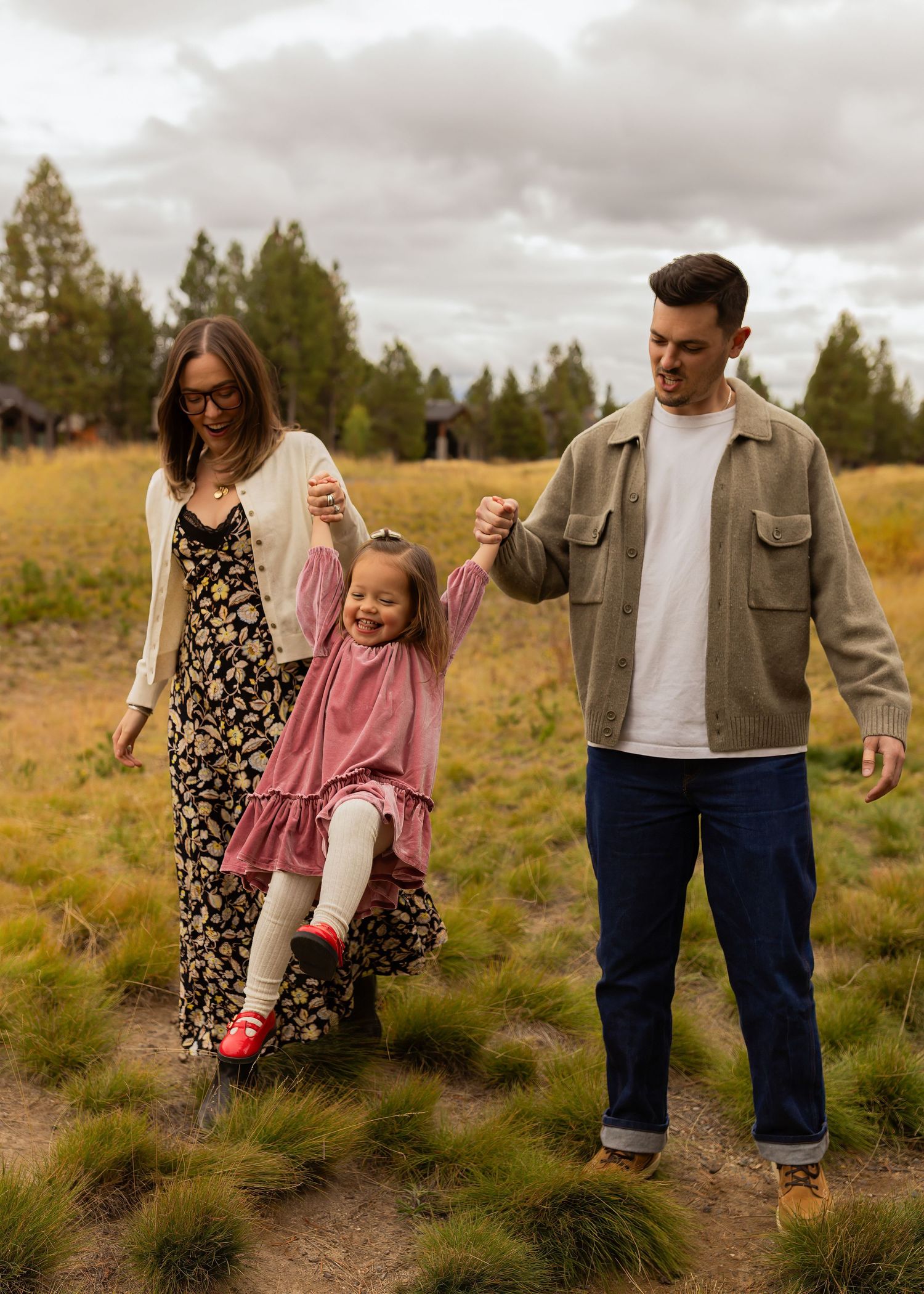 Family of three walking outdoors, parents swinging young girl between them in a meadow with evergreen trees.