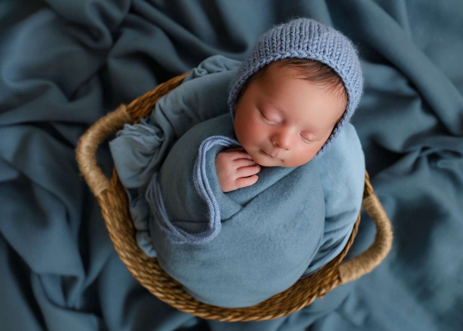 Sleeping newborn baby wrapped in blue swaddle wearing knit hat, nestled in wicker basket on blue fabric. Redmond Oregon Studio
