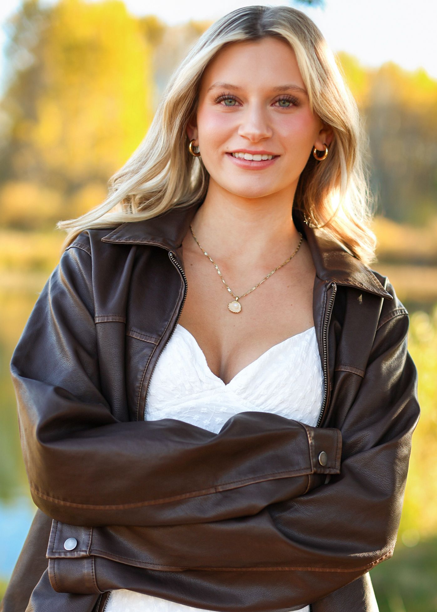 Young blonde woman in brown leather jacket and white dress, arms crossed, smiling outdoors near a lake in autumn.