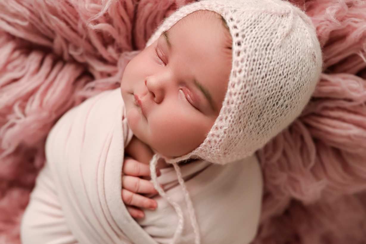 Newborn baby wearing white knit bonnet resting on pink textured fabric.
