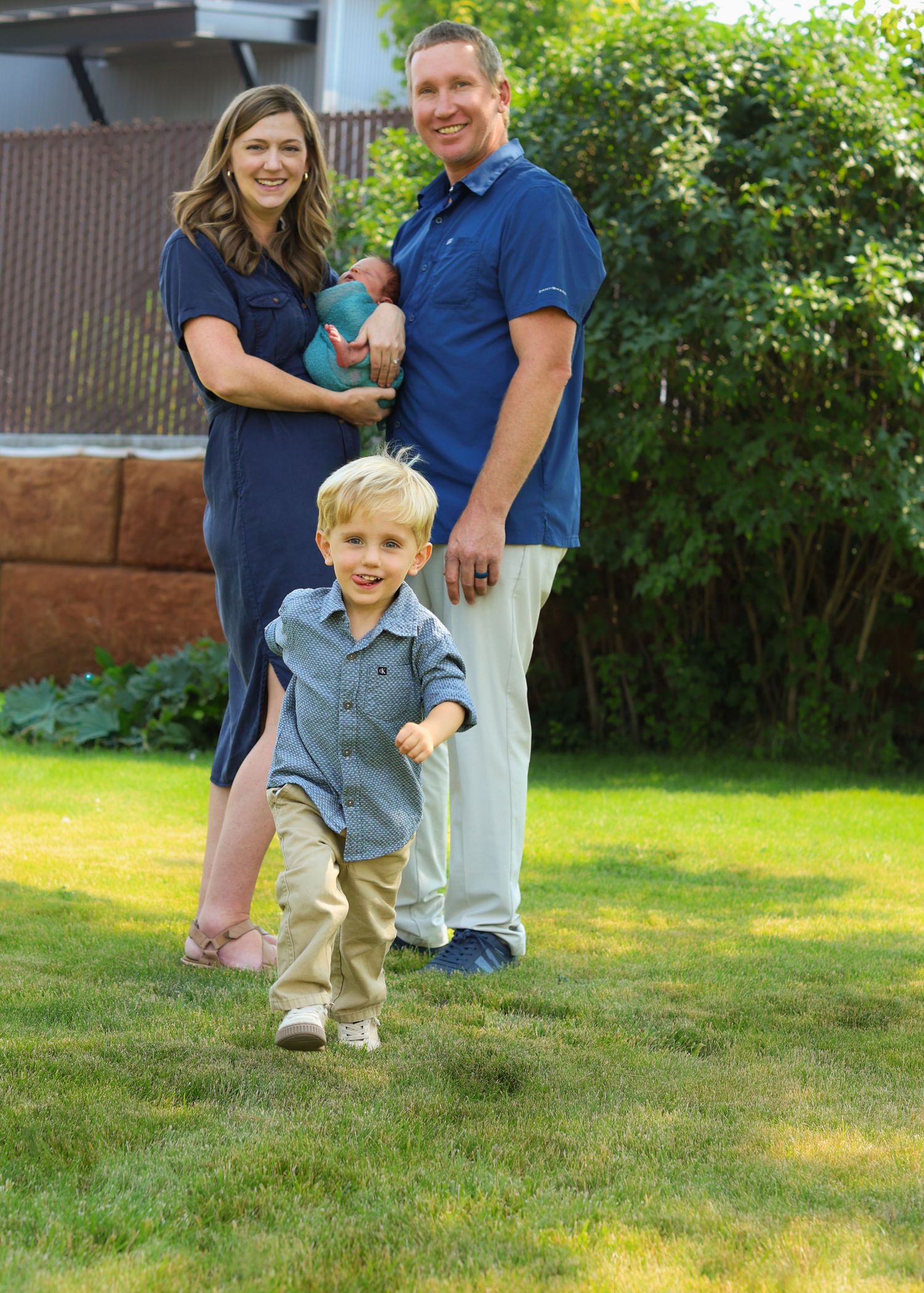 Family of four poses outdoors on green lawn, parents in navy blue holding newborn baby while toddler son runs forward.